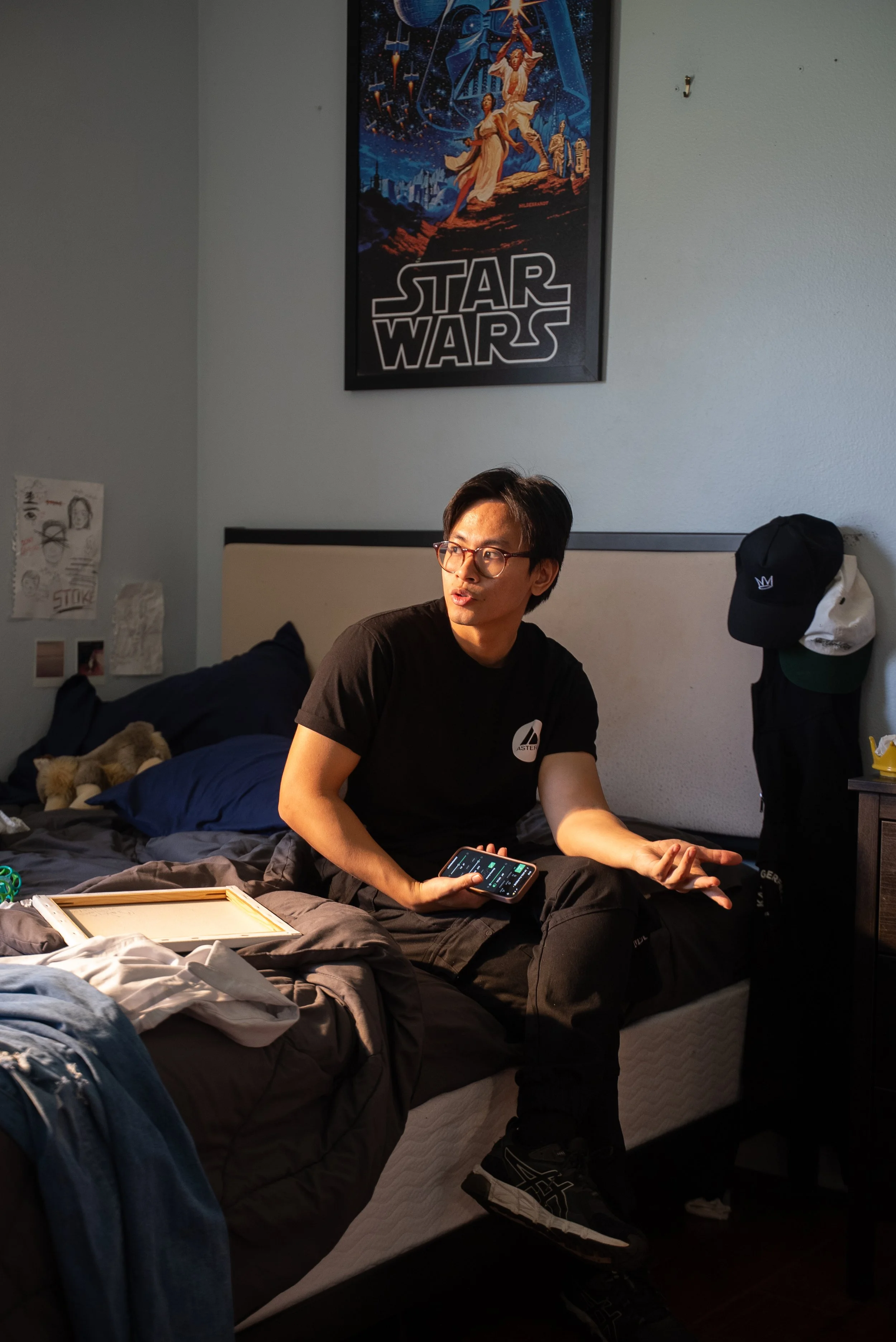 A young man with glasses and black clothing sitting on a bed in a room, holding a smartphone in his hand and gesturing with his other hand. There is a "Star Wars" poster on the wall behind him, and various items on the bed and nearby surfaces.