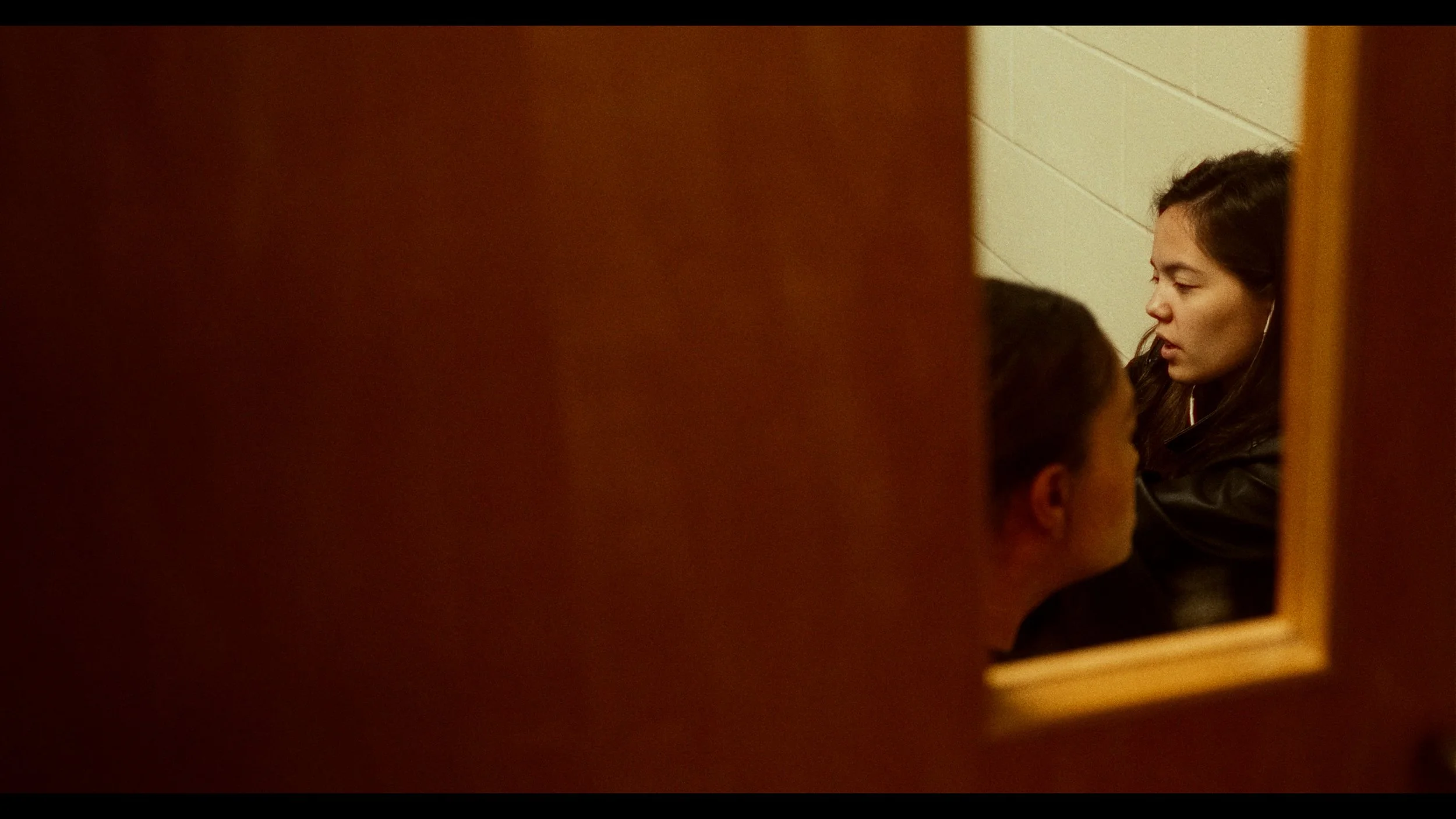 A woman with long dark hair wearing earphones is talking to a woman with short dark hair, seen from a reflection in a mirror attached to a wooden door.