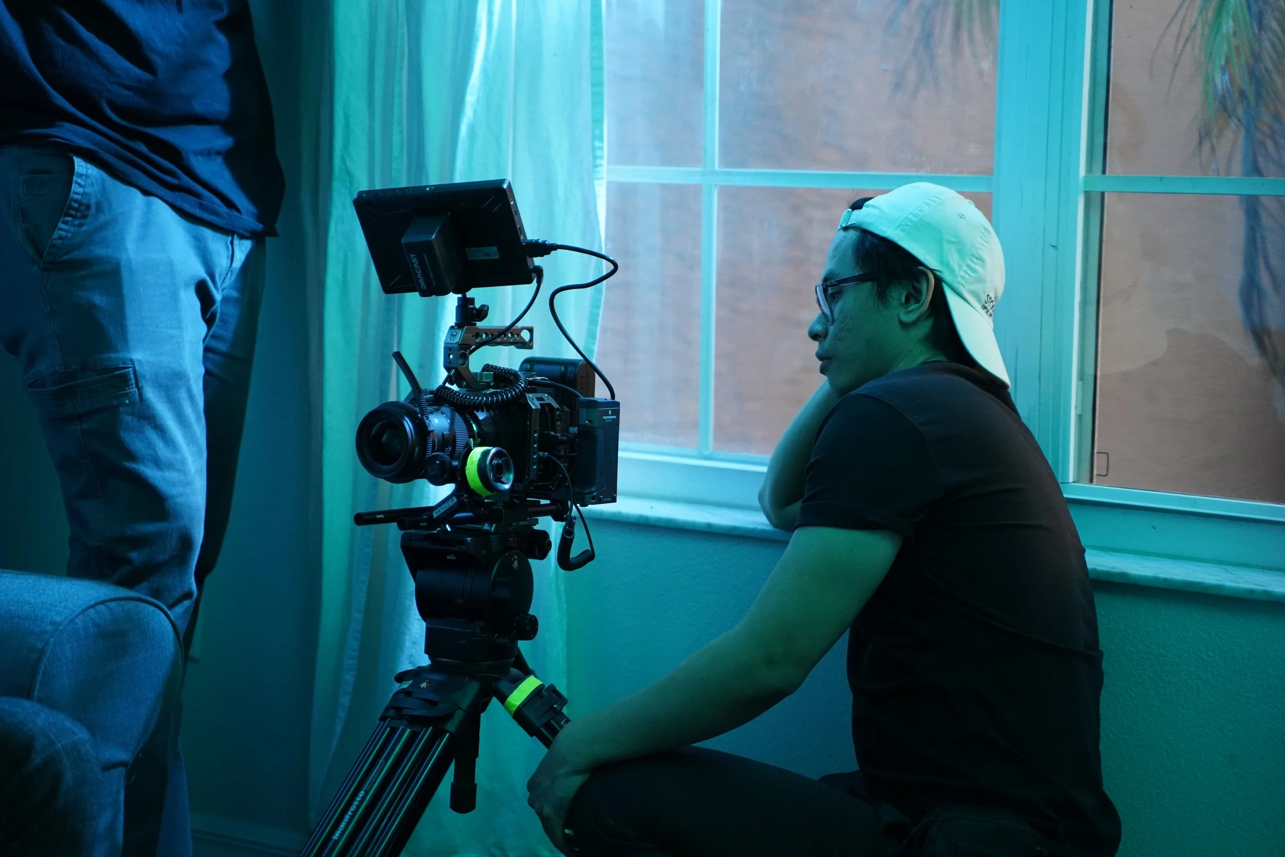 A person sitting on the floor wearing glasses and a white cap, looking at a professional film camera set up on a tripod inside a room with blue lighting.
