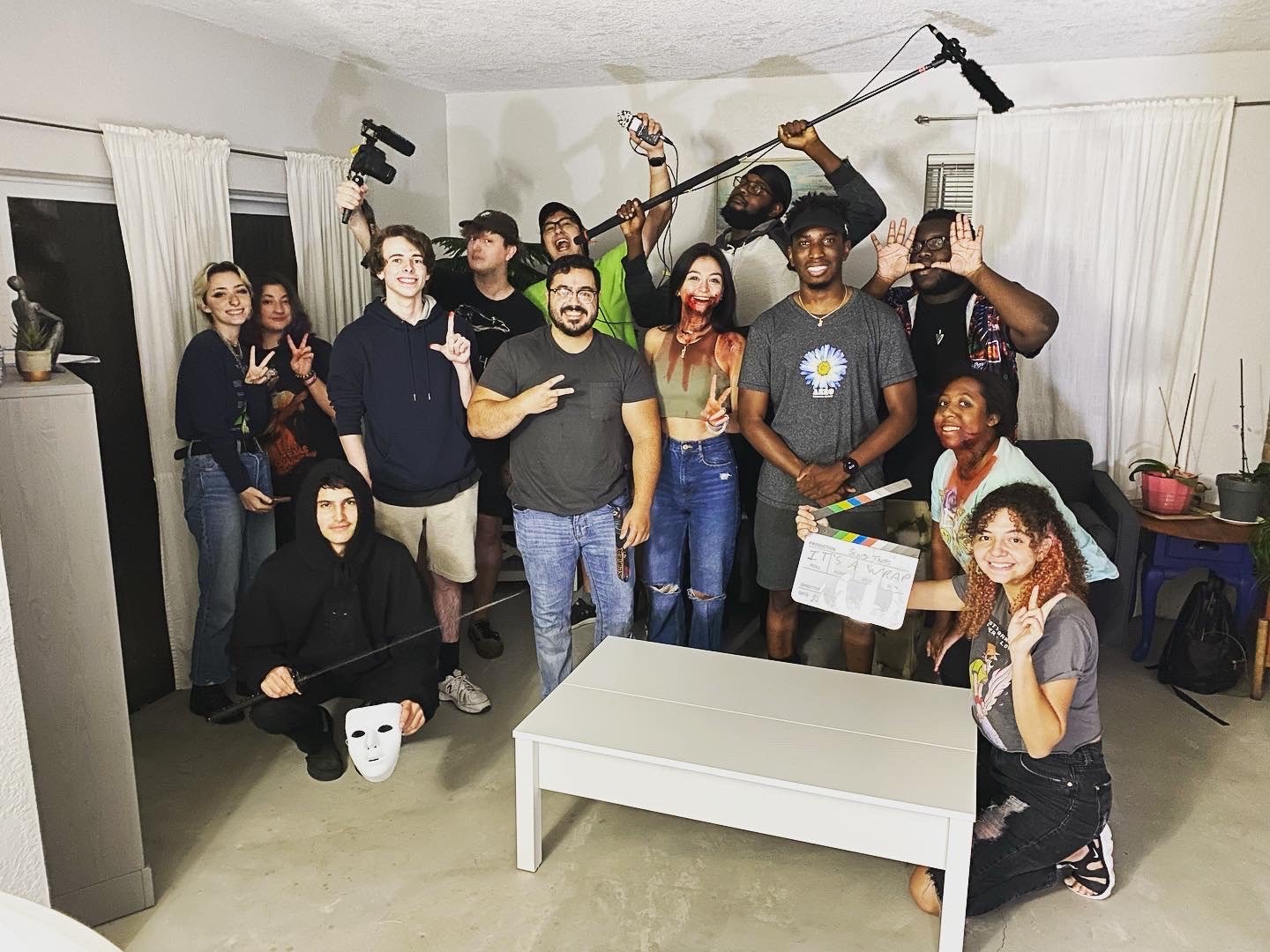 Group of people on set, including a director with a clapperboard, crew members with a boom mic, and cast members dressed as horror characters, celebrating after filming.