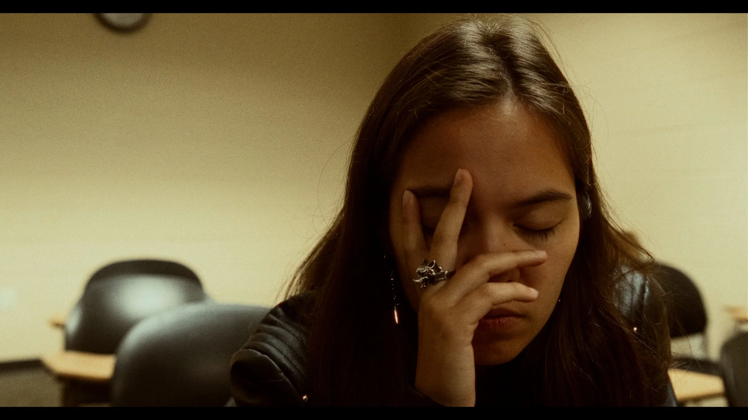 Young woman in a classroom with closed eyes, holding her forehead with her fingers, wearing rings and a black leather jacket.