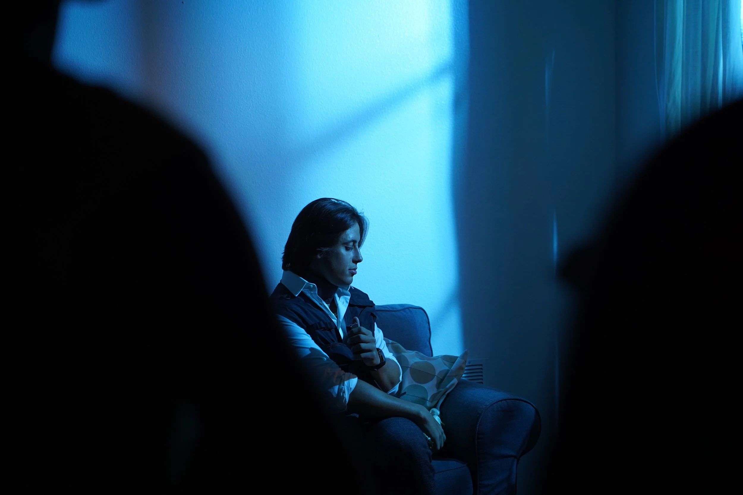 A man sitting on a blue sofa in a dimly lit room with blue lighting, framed by dark silhouettes on either side.