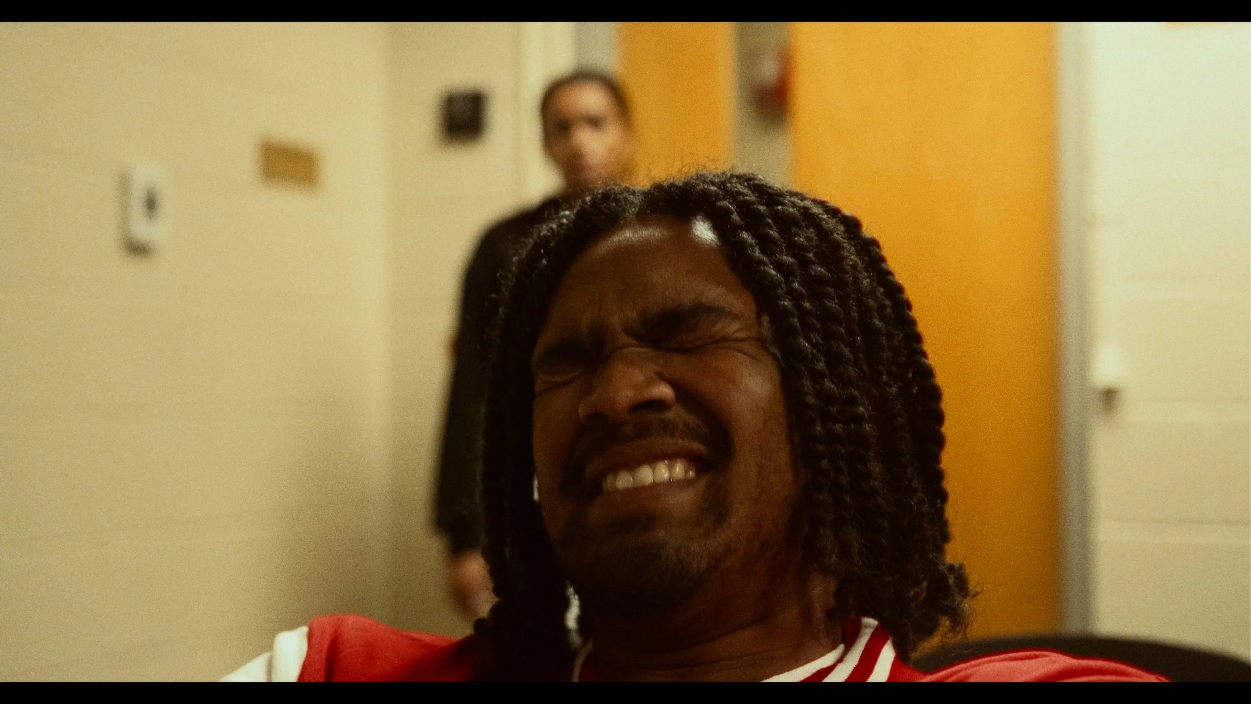 A man with long dreadlocks making a facial expression of pain or discomfort, eyes closed tightly, mouth open showing teeth, sitting indoors with a person in the background.