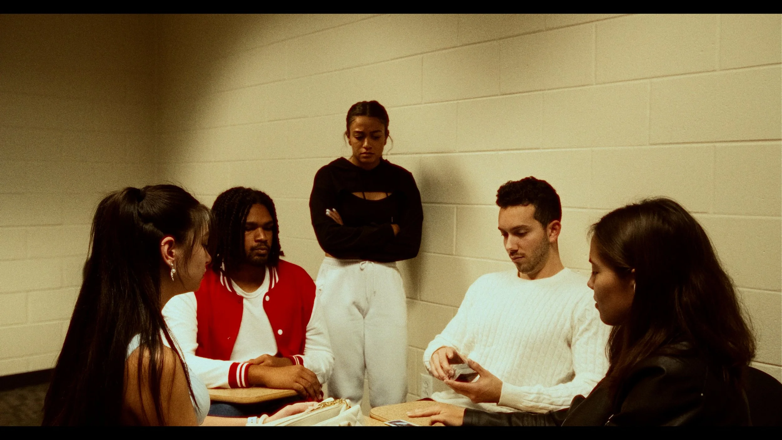 Five teenagers in a classroom or study room, with three seated around a table and two standing, looking serious or contemplative.