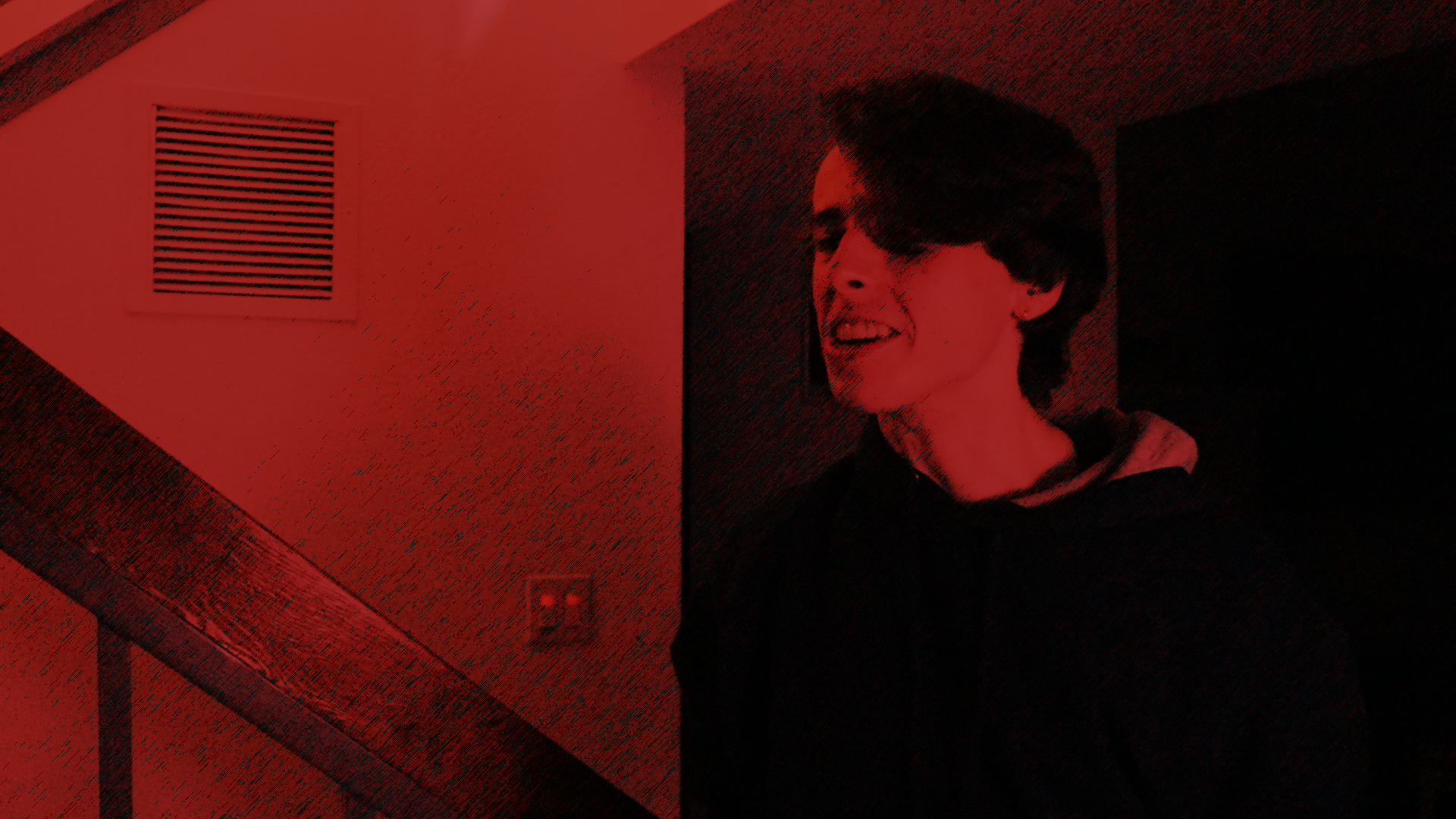 A young man with dark hair and glasses smiling, wearing a hoodie, standing in a dimly lit room with red lighting, a vent on the wall, and a staircase.