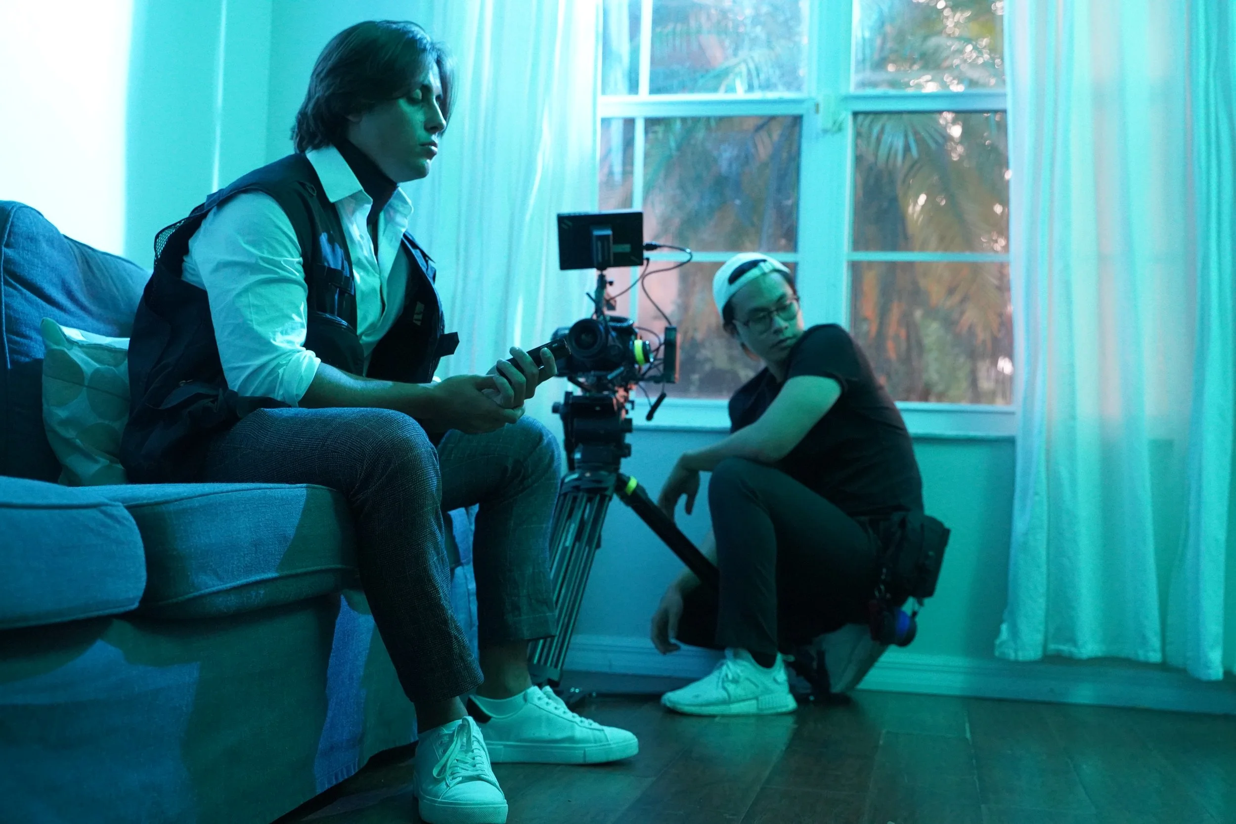 Two young men on a film set, one sitting on a couch holding a camera and the other crouching beside a camera on a tripod in a room with a large window and blue lighting.