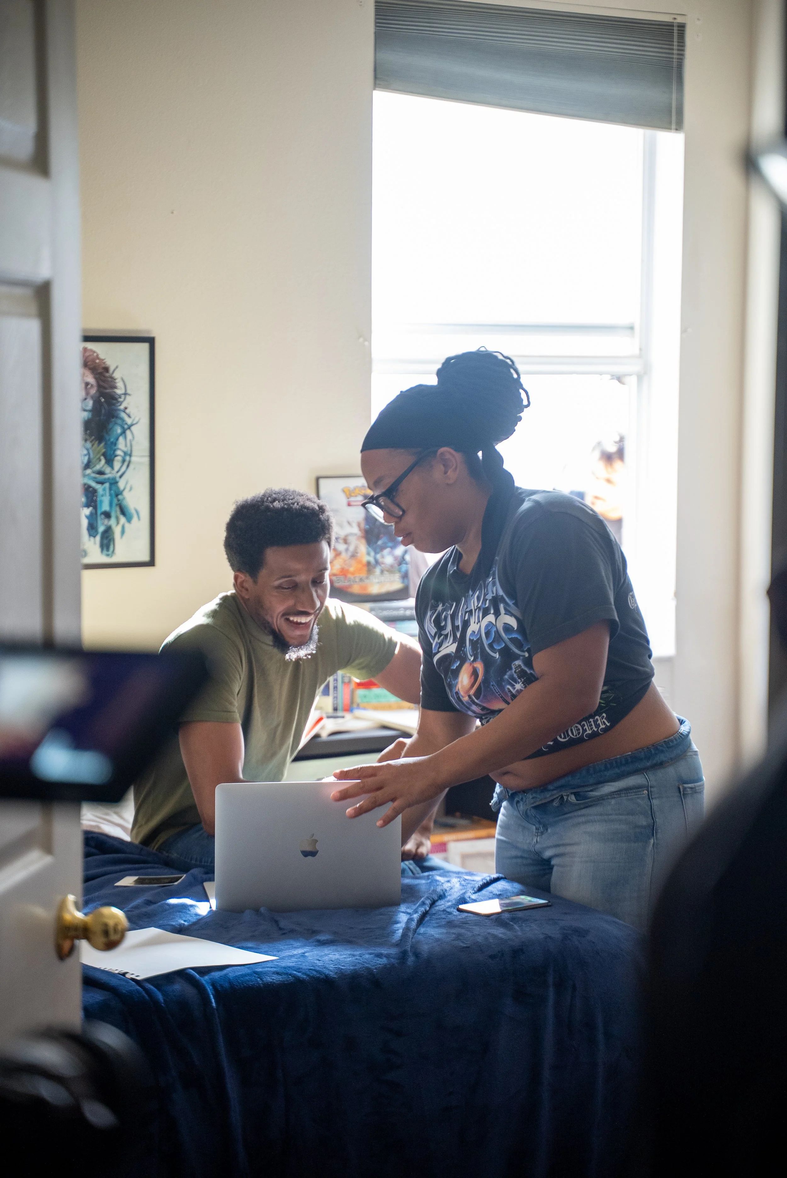 Two people in a bedroom, one sitting on the bed and the other leaning over, looking at a laptop with smiles, surrounded by posters and books, with a window in the background.