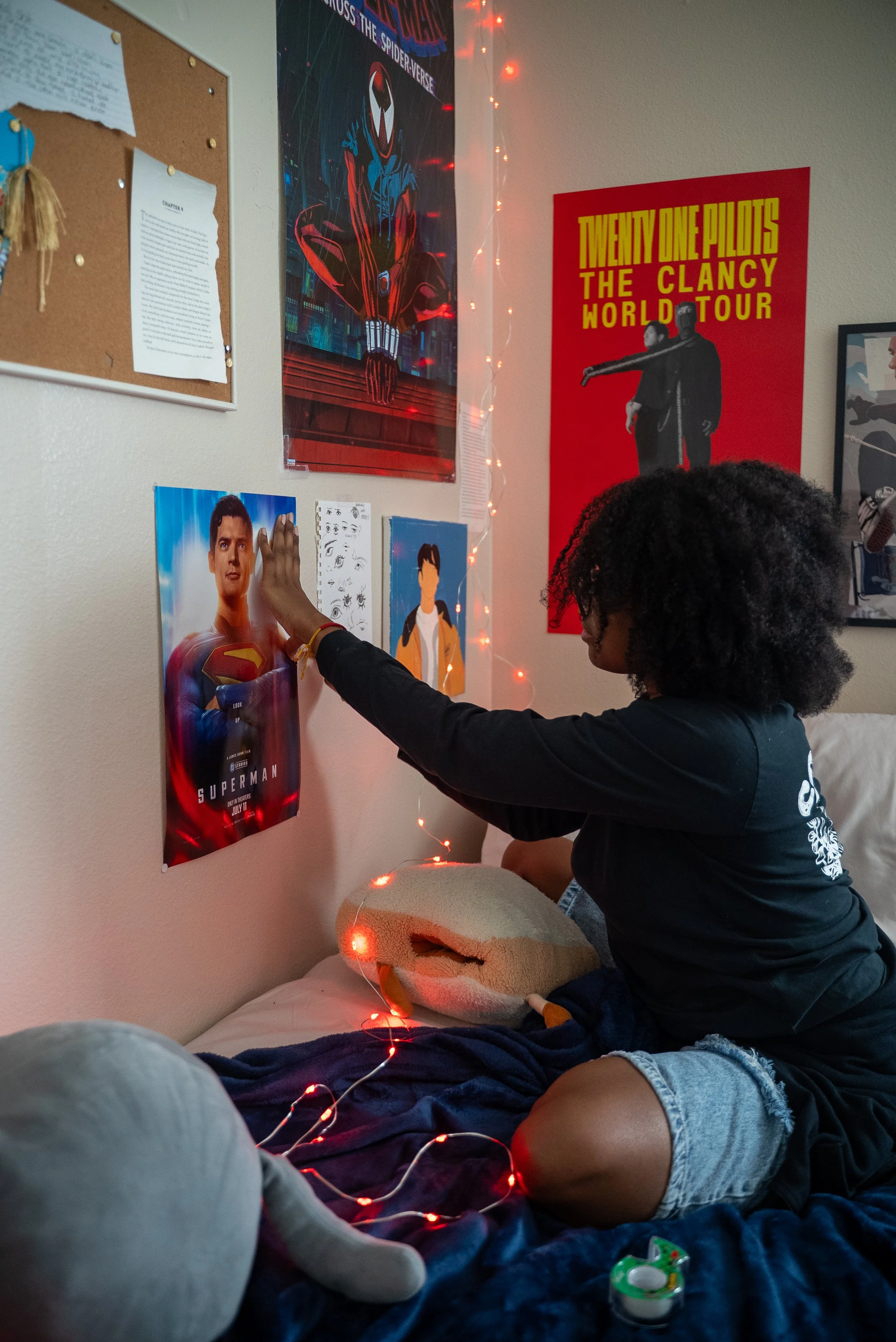 A person sitting on a bed decorated with string lights, hanging posters of superhero and music band on the wall, and touching a Superman poster, with plush toys and a pacifier on the bed.