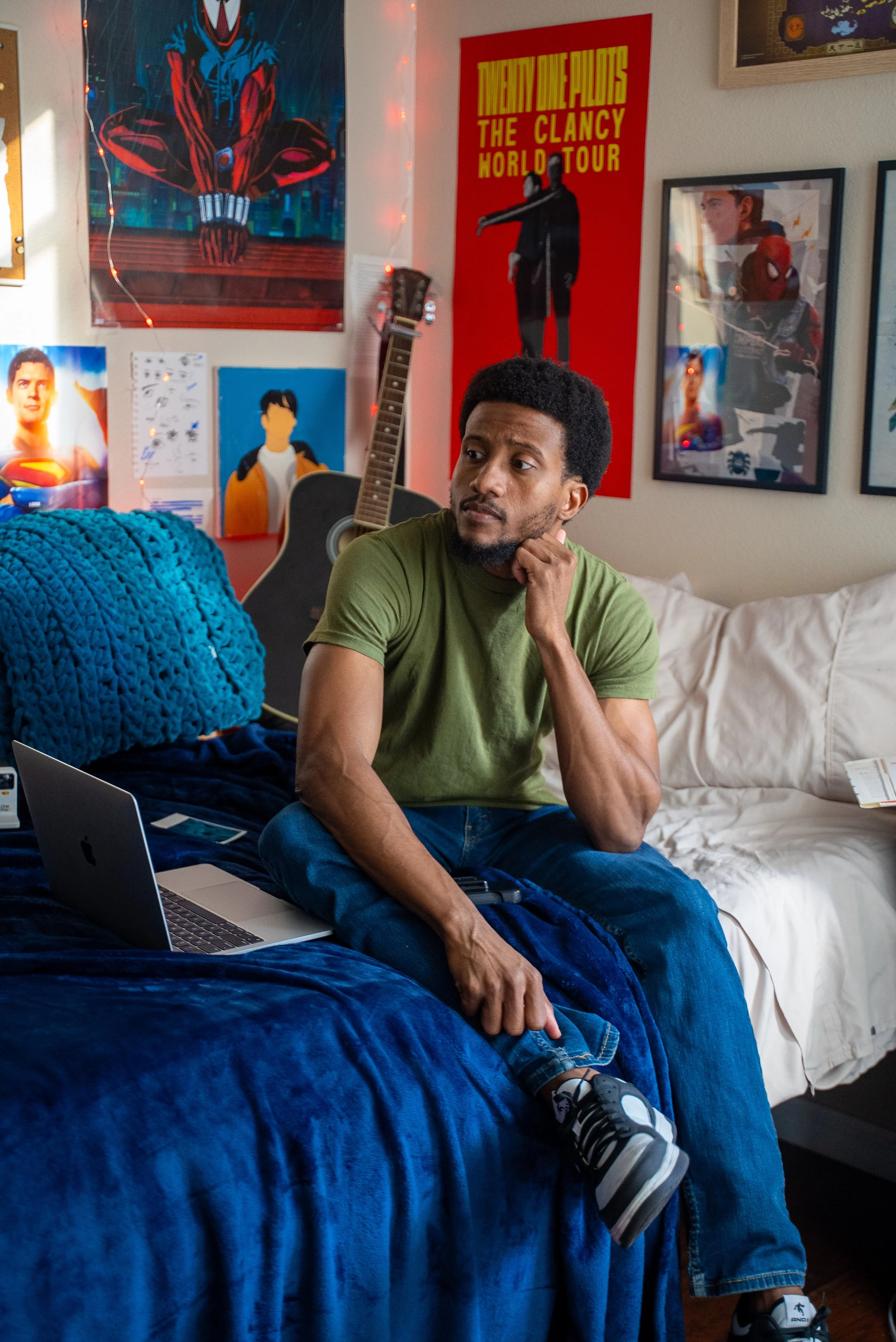 A young man sitting on a bed in a room decorated with superhero posters and artwork, listening thoughtfully to something on his phone. There's a laptop open on the bed, and a guitar leaning against the wall behind him.