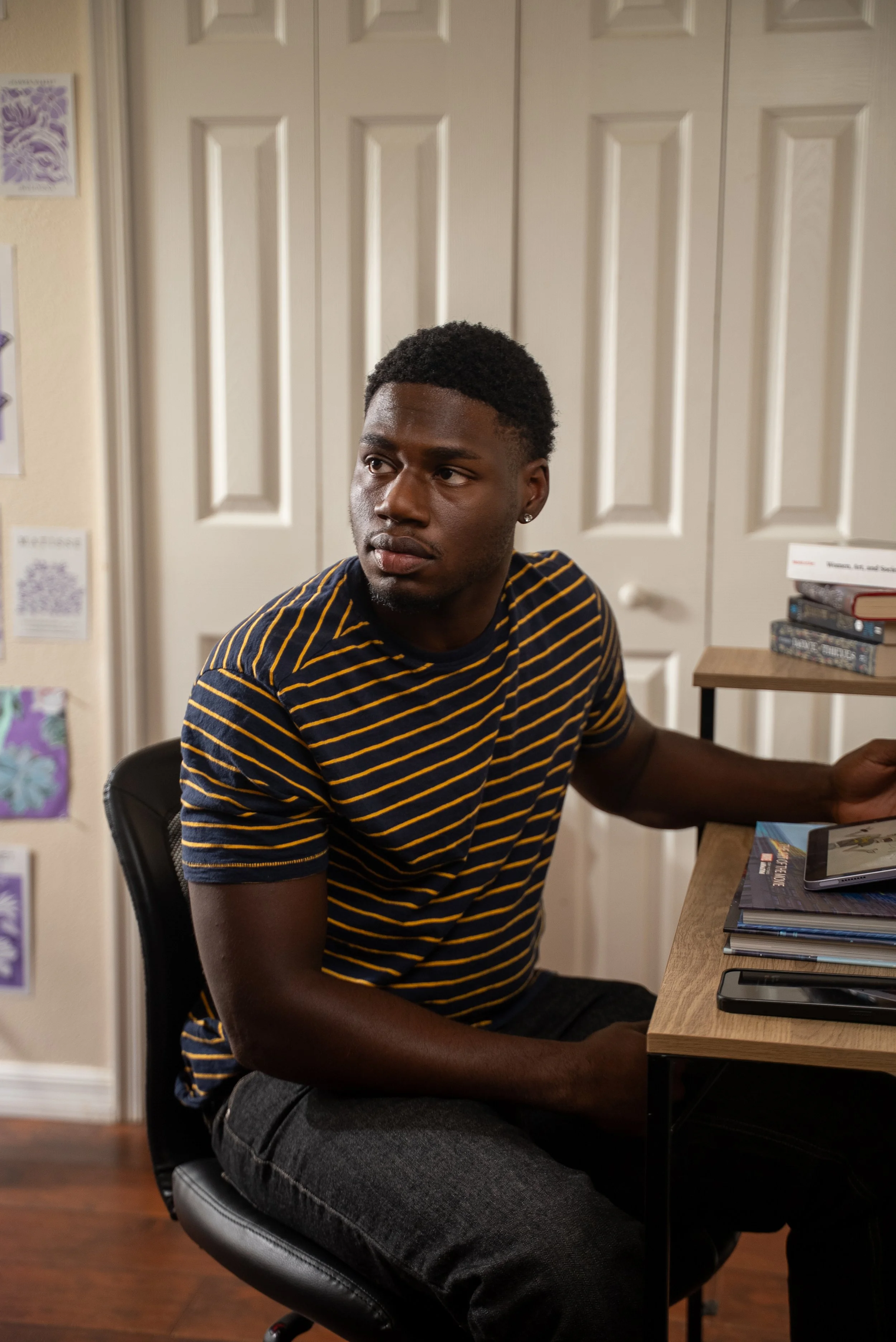 A young man with dark skin, short curly hair, and a navy and yellow striped shirt sitting at a wooden desk in a room, looking to his left. The background features white closet doors and some books on a nearby shelf.