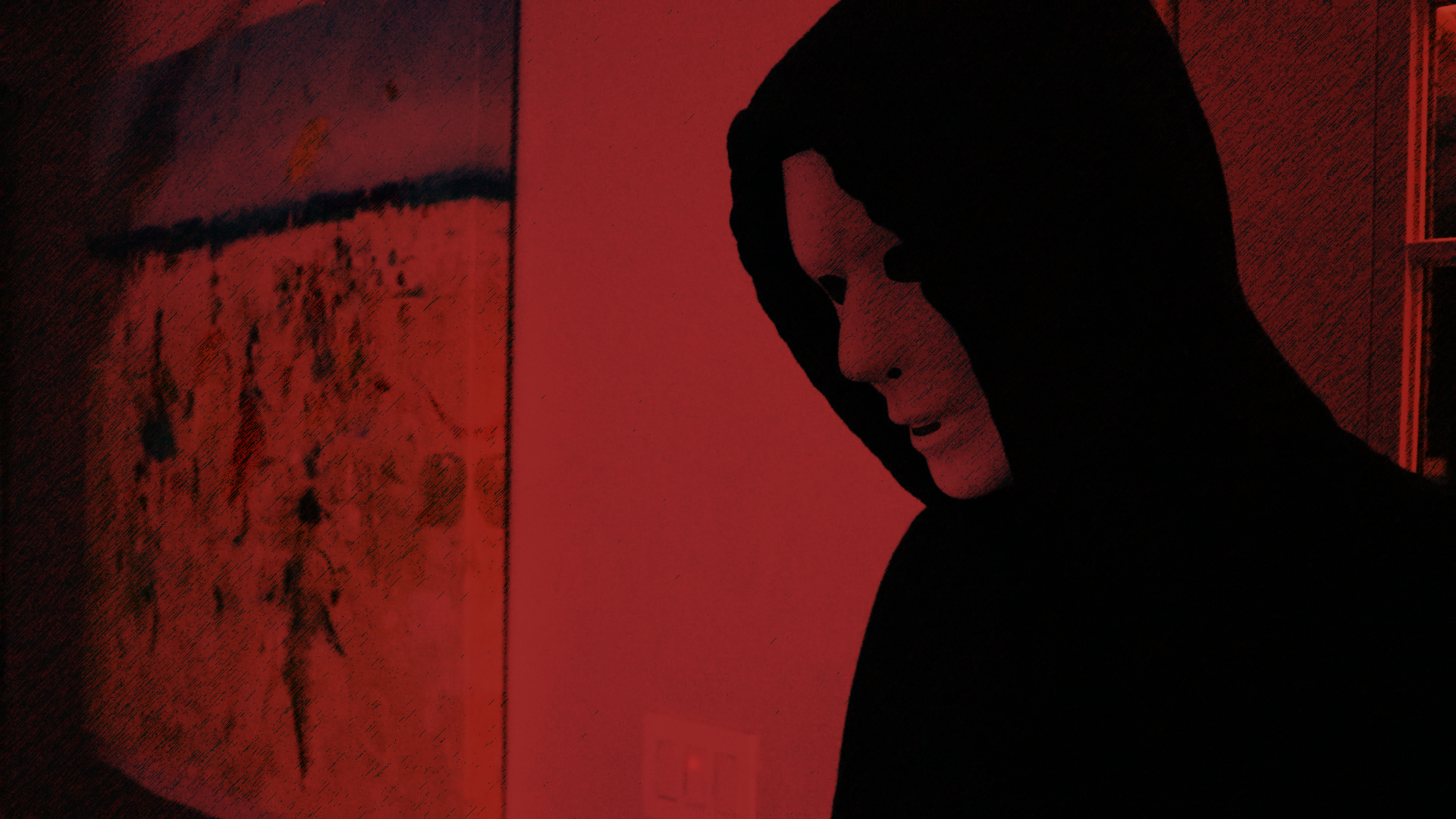 A person wearing a black hoodie looking down in a room with dark red lighting.