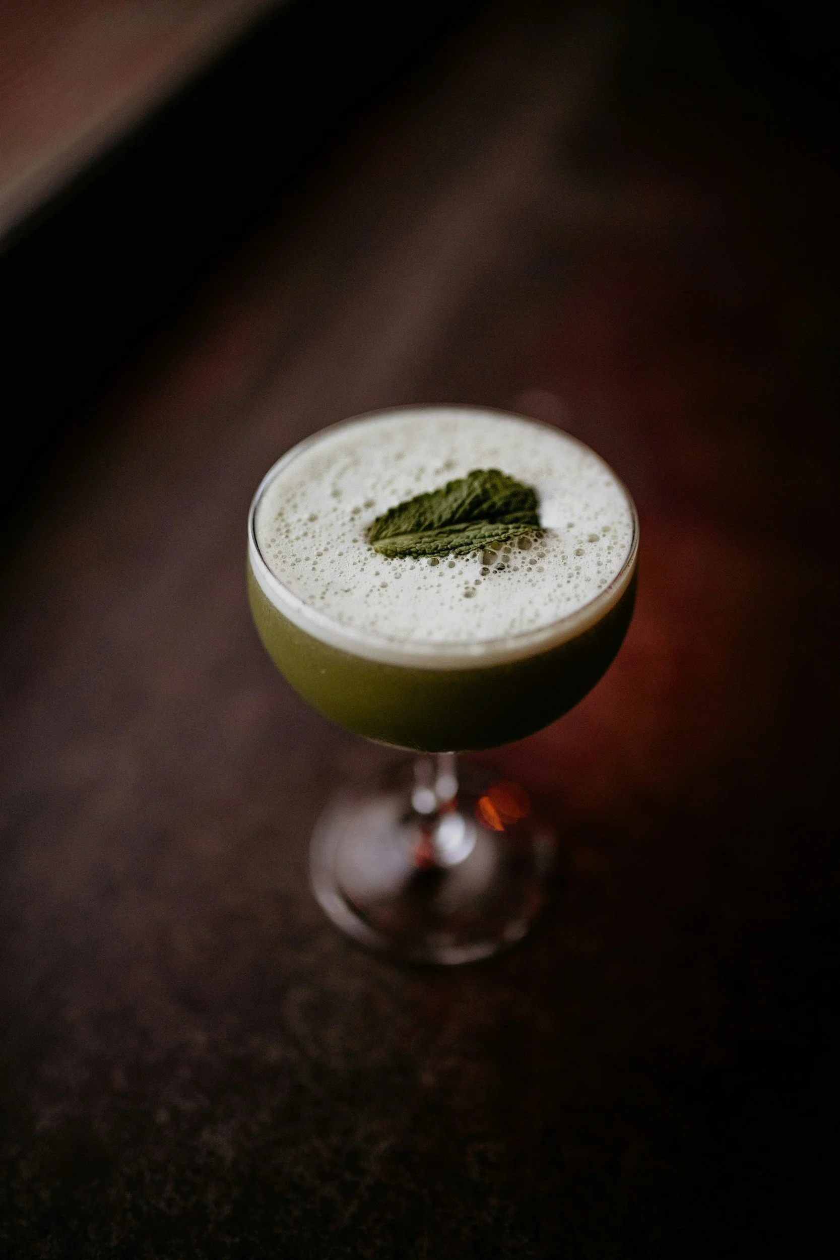 A cocktail in a stemmed glass with a foam top and two mint leaves as garnish on a dark surface.