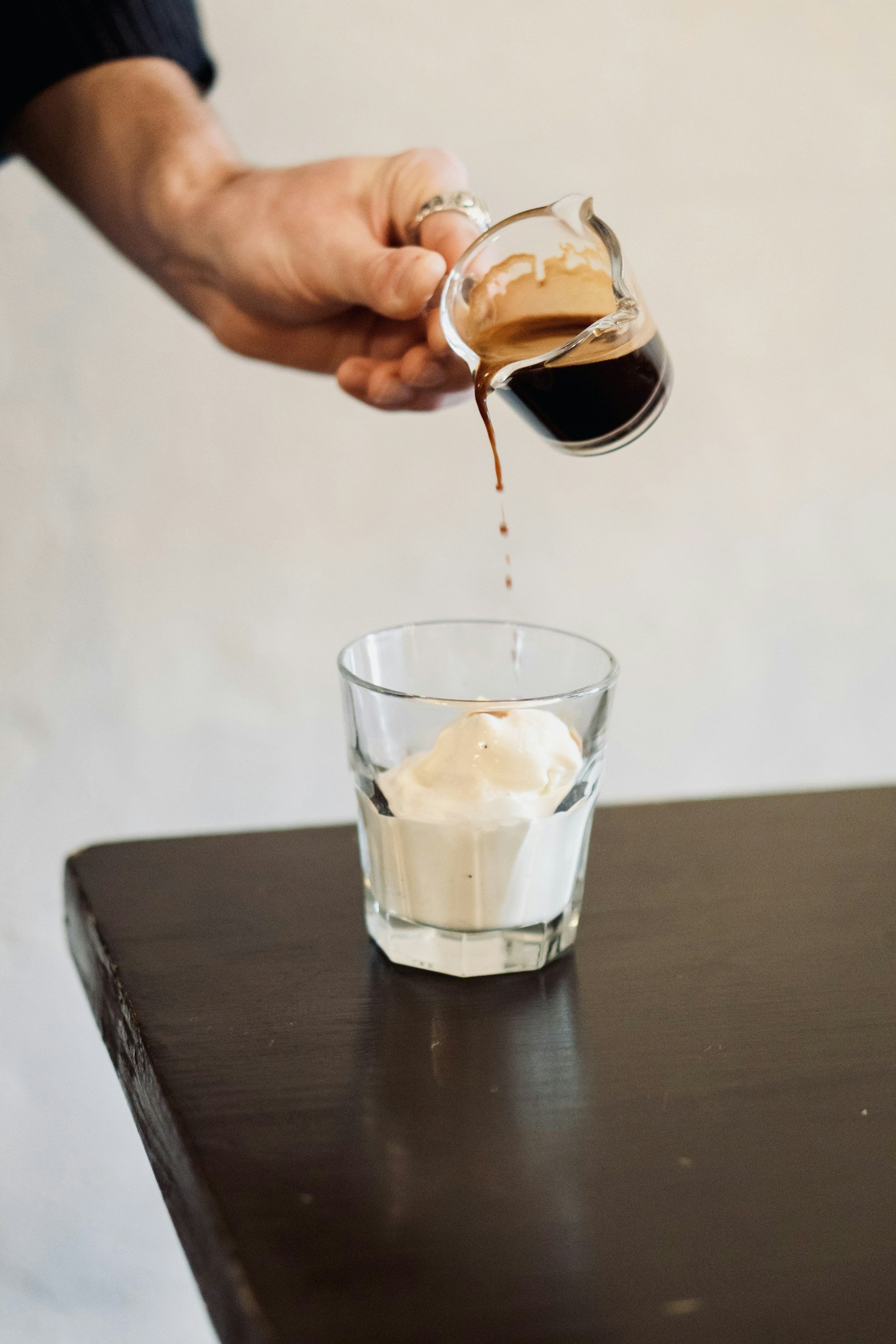 A person holds a small glass pitcher filled with dark coffee, pouring it into a tall glass with a scoop of vanilla ice cream, on a dark wooden table.