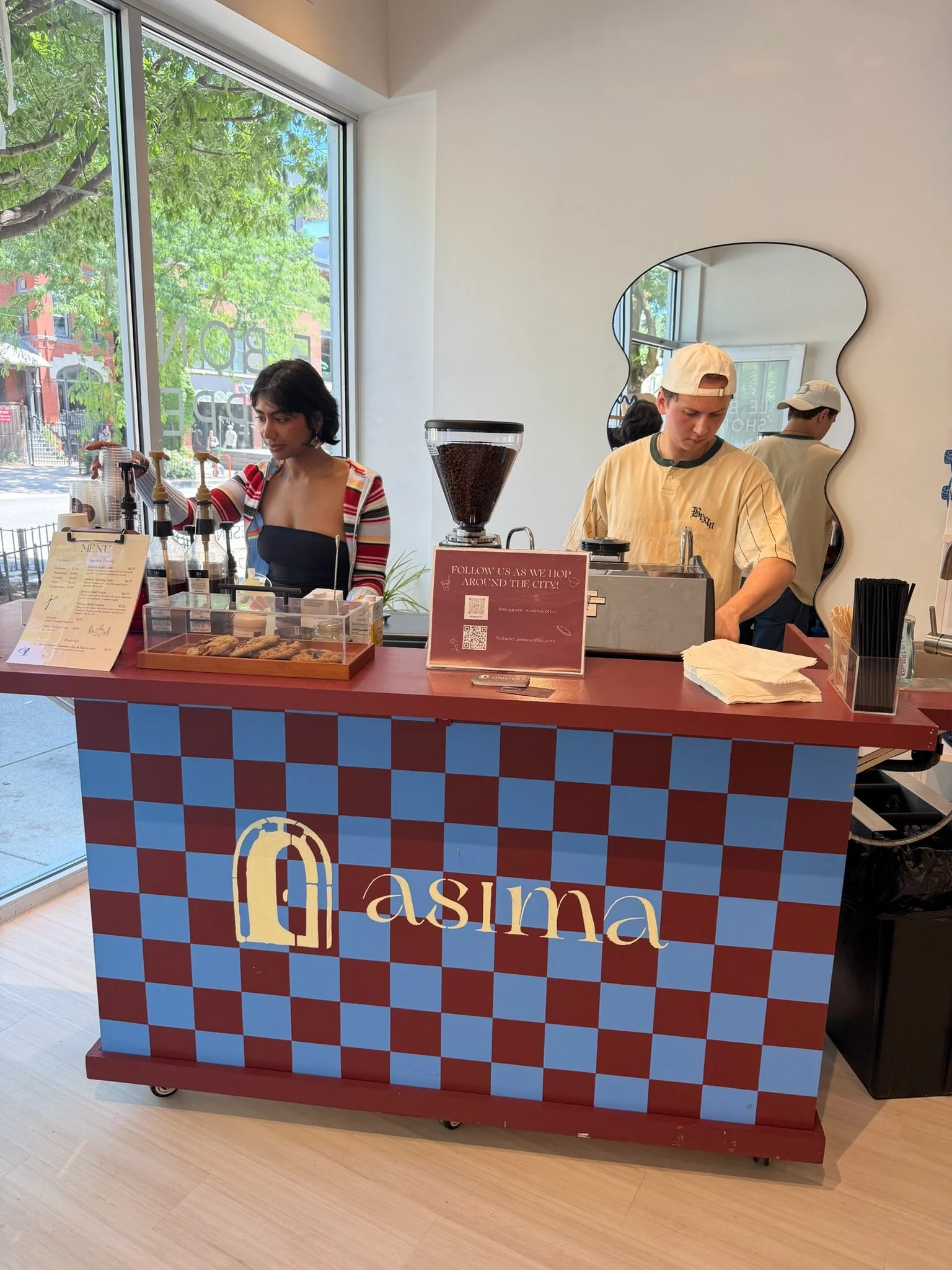 A coffee counter with a checkered pink and blue pattern and the word 'asia' on the front. Two baristas are behind the counter, one woman and one man, preparing coffee. A large window behind them reveals trees and buildings outside.
