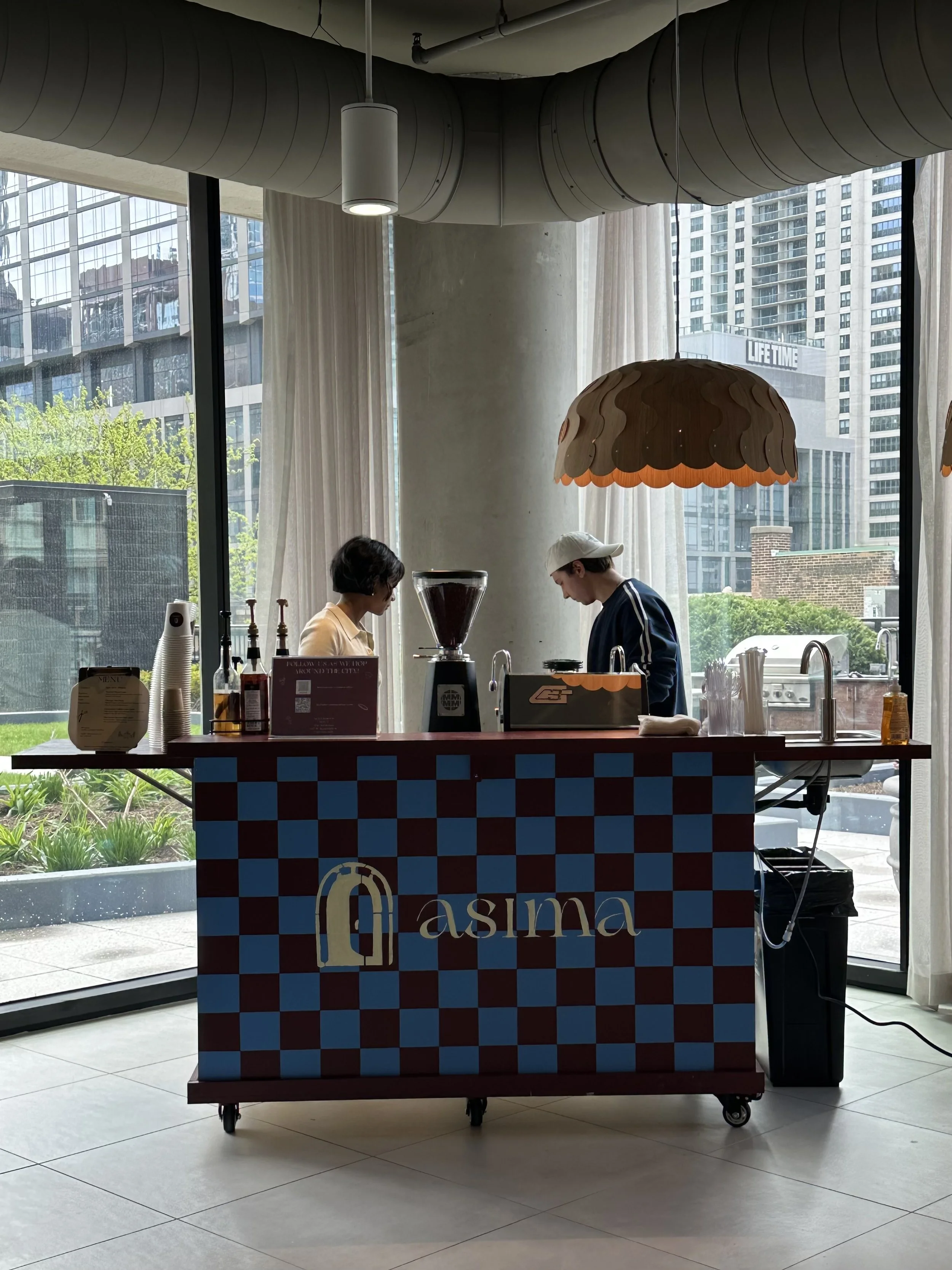 Indoor scene with a coffee stand labeled 'Qasima' in front of large windows showing city buildings. Two baristas are preparing drinks; one is wearing glasses, and the other is wearing a cap. The stand has a checkered pattern and is equipped with coffee brewing equipment, cups, and supplies. Overhead, there are modern hanging light fixtures and large ventilation ducts.