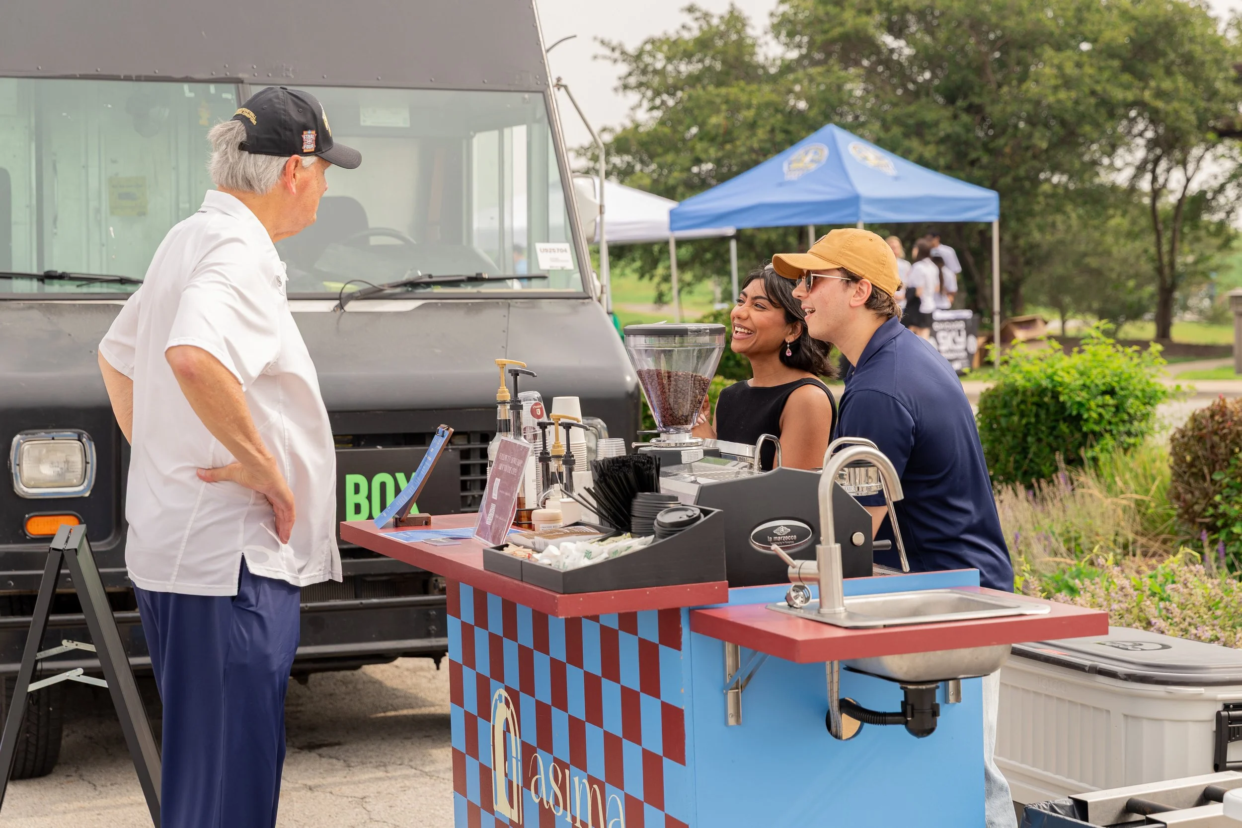A man in a white shirt and black cap talks to two young adults, a woman and a man, at a food stand with a red and blue checkered pattern. The stand has coffee beans, cups, and a beverage dispenser. Behind them, there is a food truck and green trees, indicating an outdoor event.