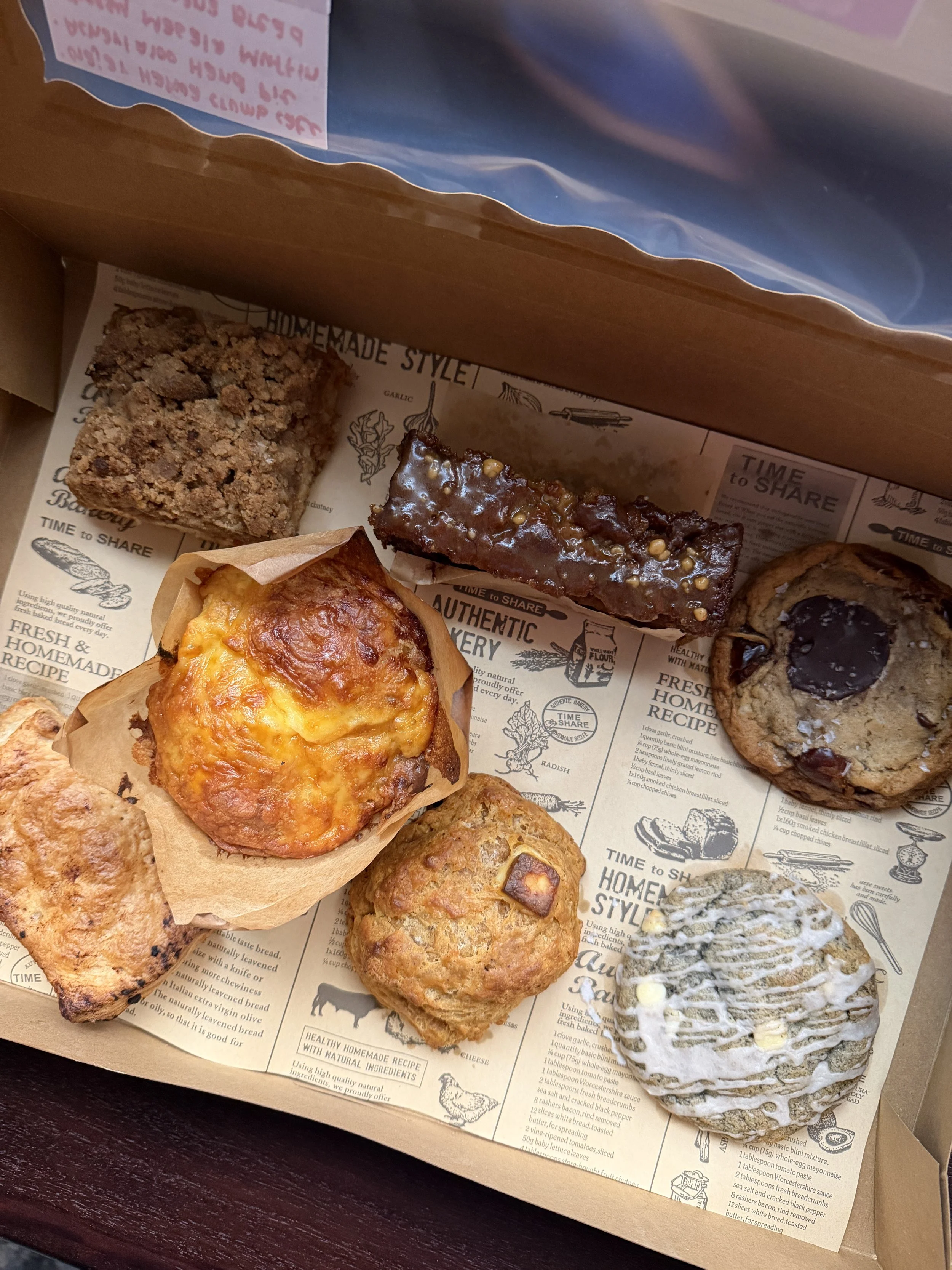 Assorted baked goods including cookies, a muffin, and a decorated cake on a paper lining in a bakery box.