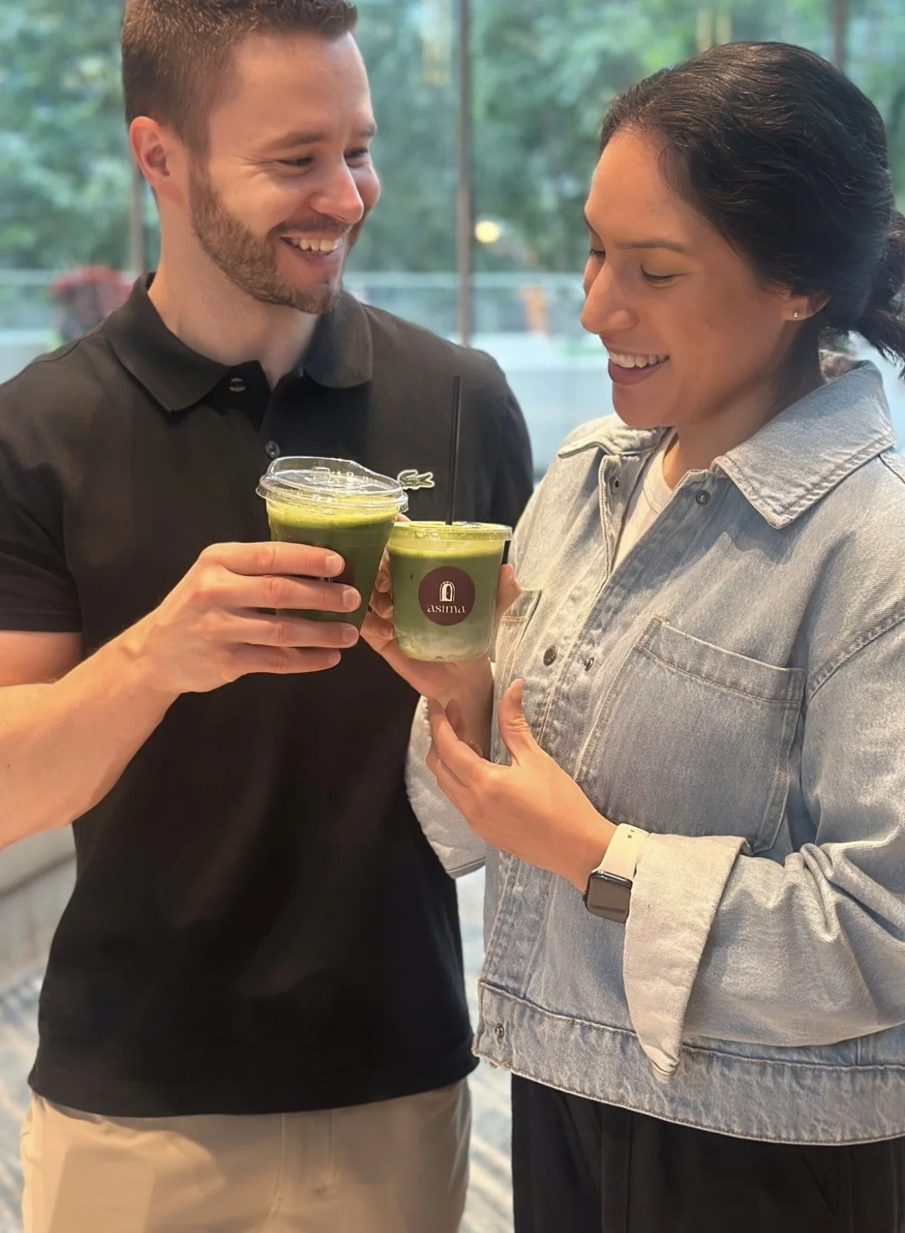 Two people are smiling and holding green smoothies together indoors, with a window showing trees outside.