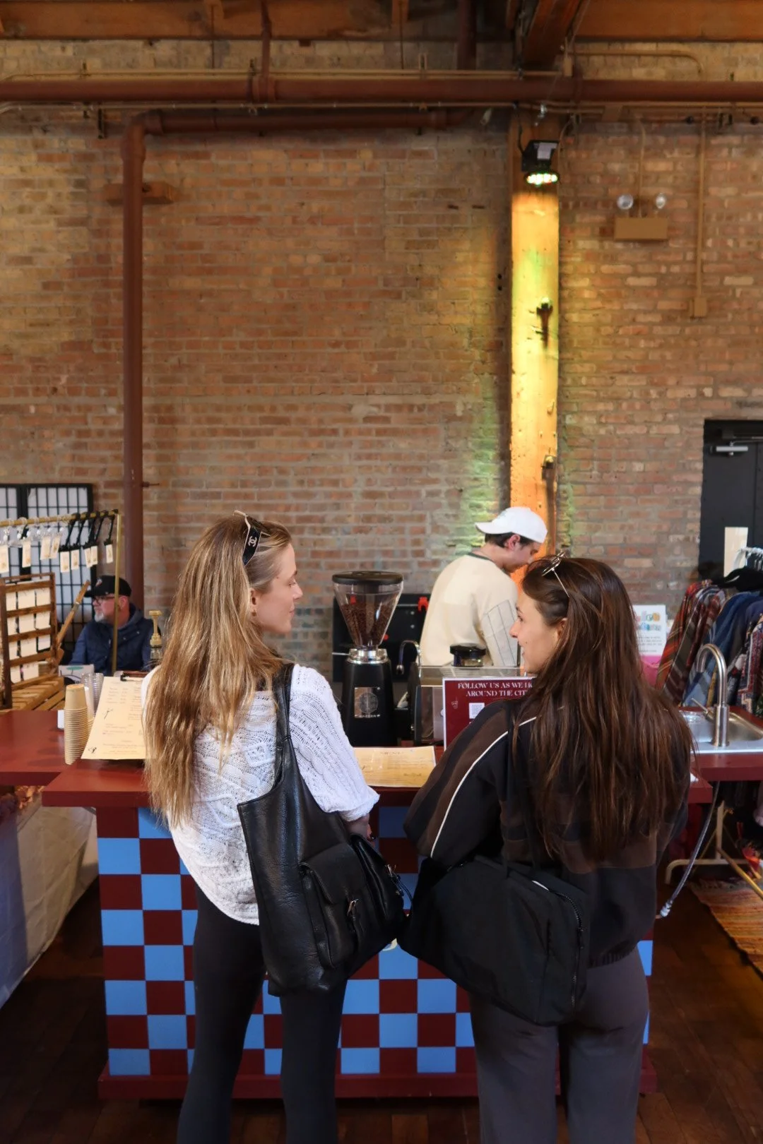 Two women with long hair and backpacks standing at a coffee shop counter, talking, with a barista working behind the counter and brick wall background.