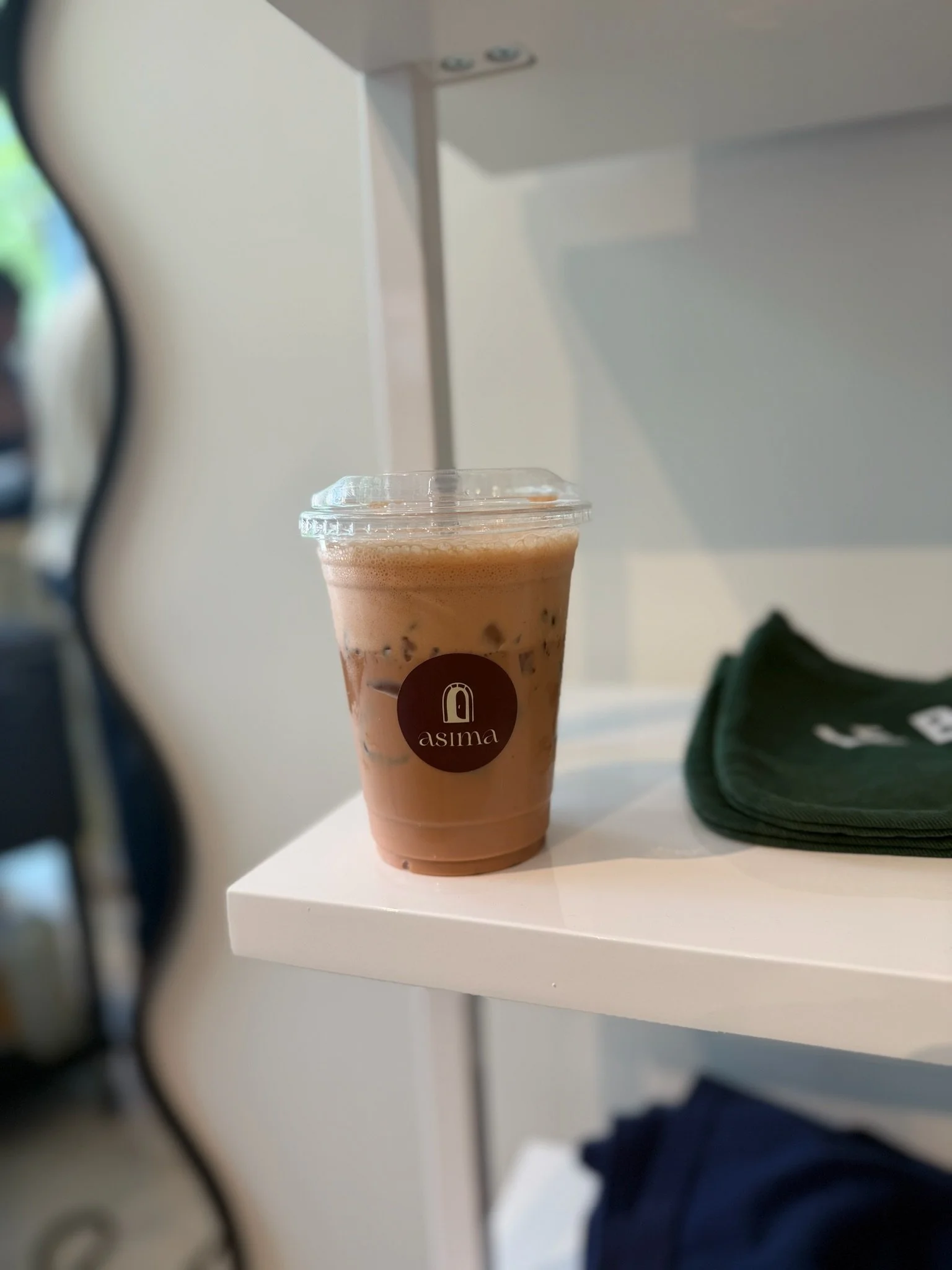 Close-up of a plastic cup filled with iced coffee or tea, with a black straw and a brown round sticker with the word 'asima' on it, placed on a white table in an indoor setting.