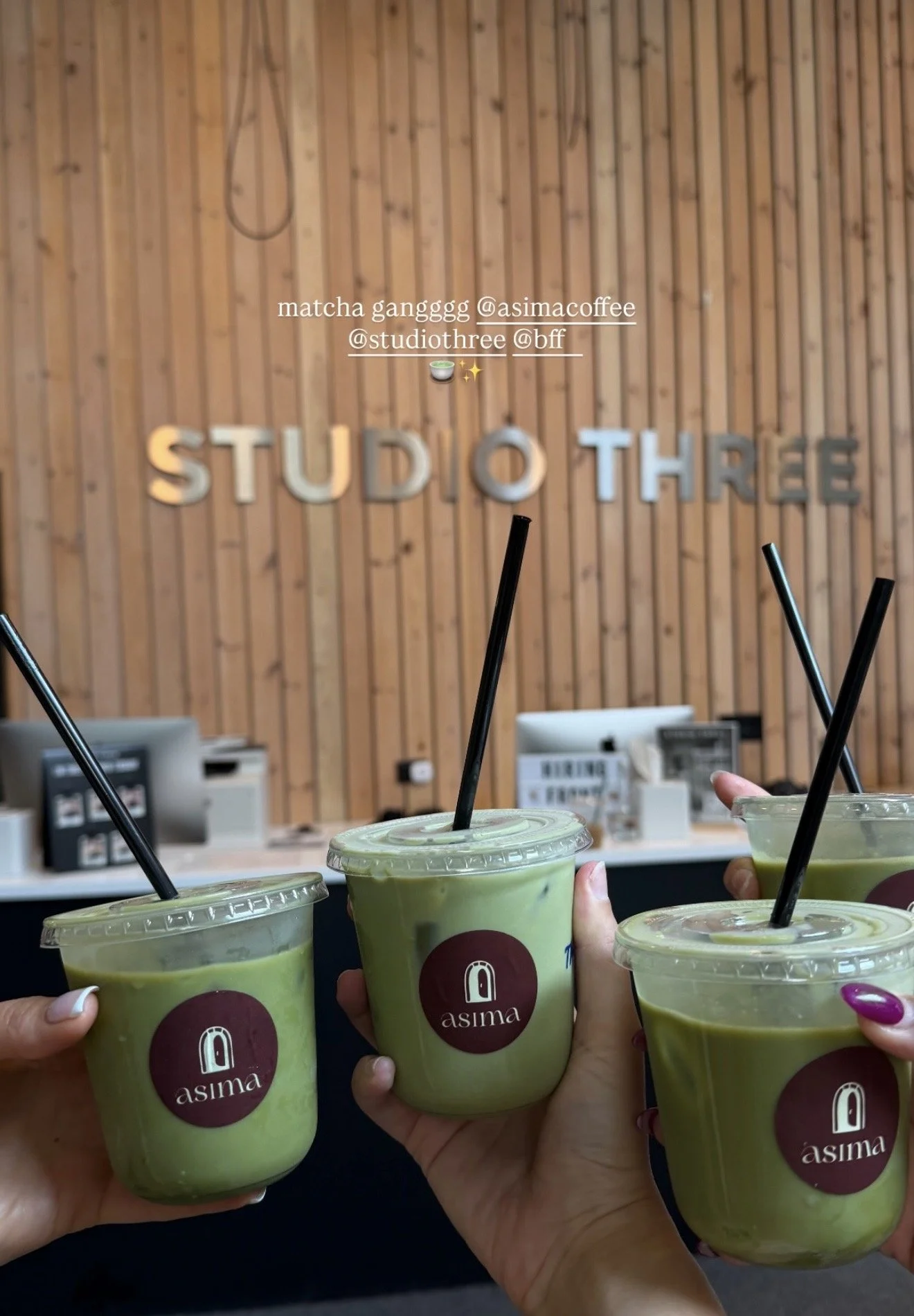 Several hands holding cups of green matcha latte at a cafe named Asima, with a wooden panel wall and signage for Studio Three in the background.