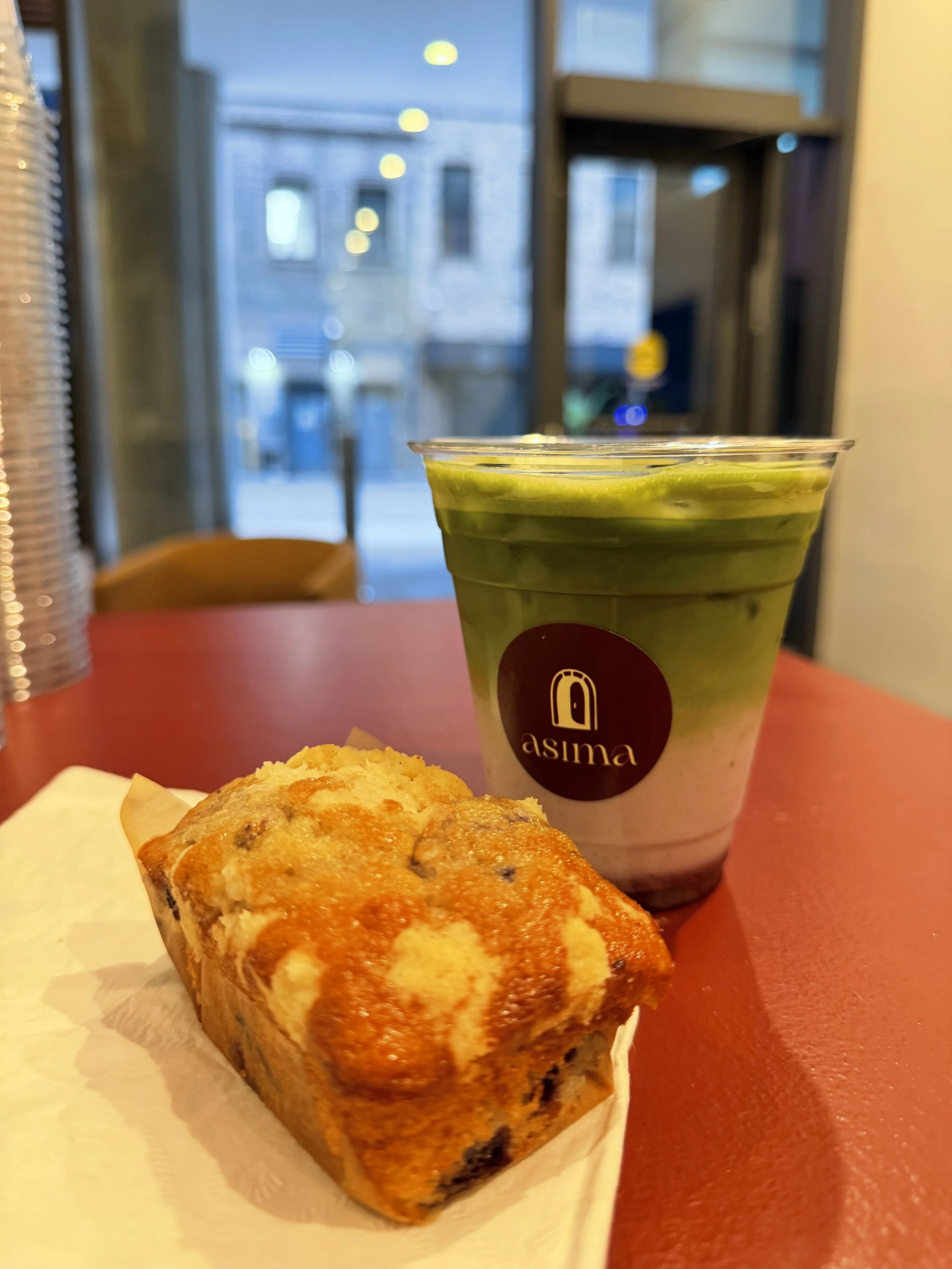 Close-up of a slice of banana bread with chocolate chips, a green matcha latte in a clear plastic cup with a red logo, and a yellow decorative item on a red table in a cafe setting, with blurred window and building outside.