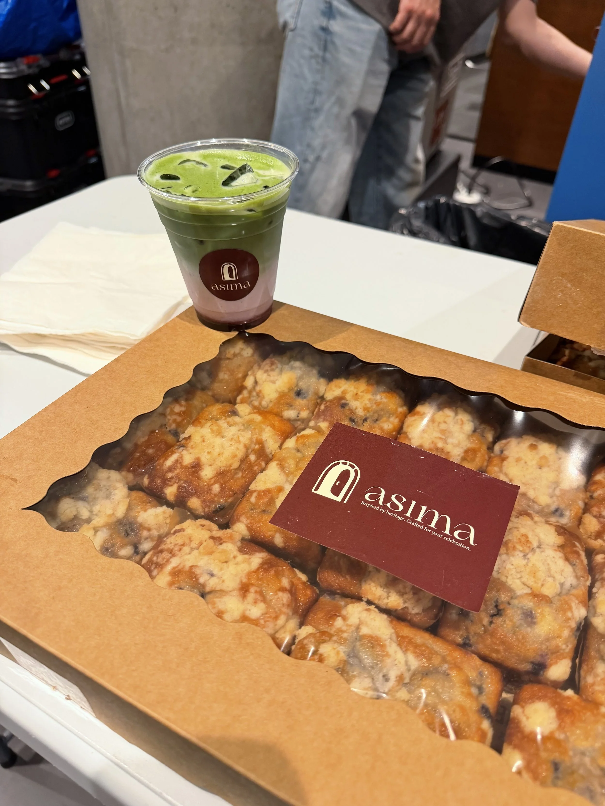 A box of sliced pepperoni pizza with melted cheese on a cardboard tray, with a sticker that says 'asima' and a logo, on a white surface. In the background, a cup of green iced matcha latte with ice cubes, a logo that says 'asima', and a person wearing gray jeans.