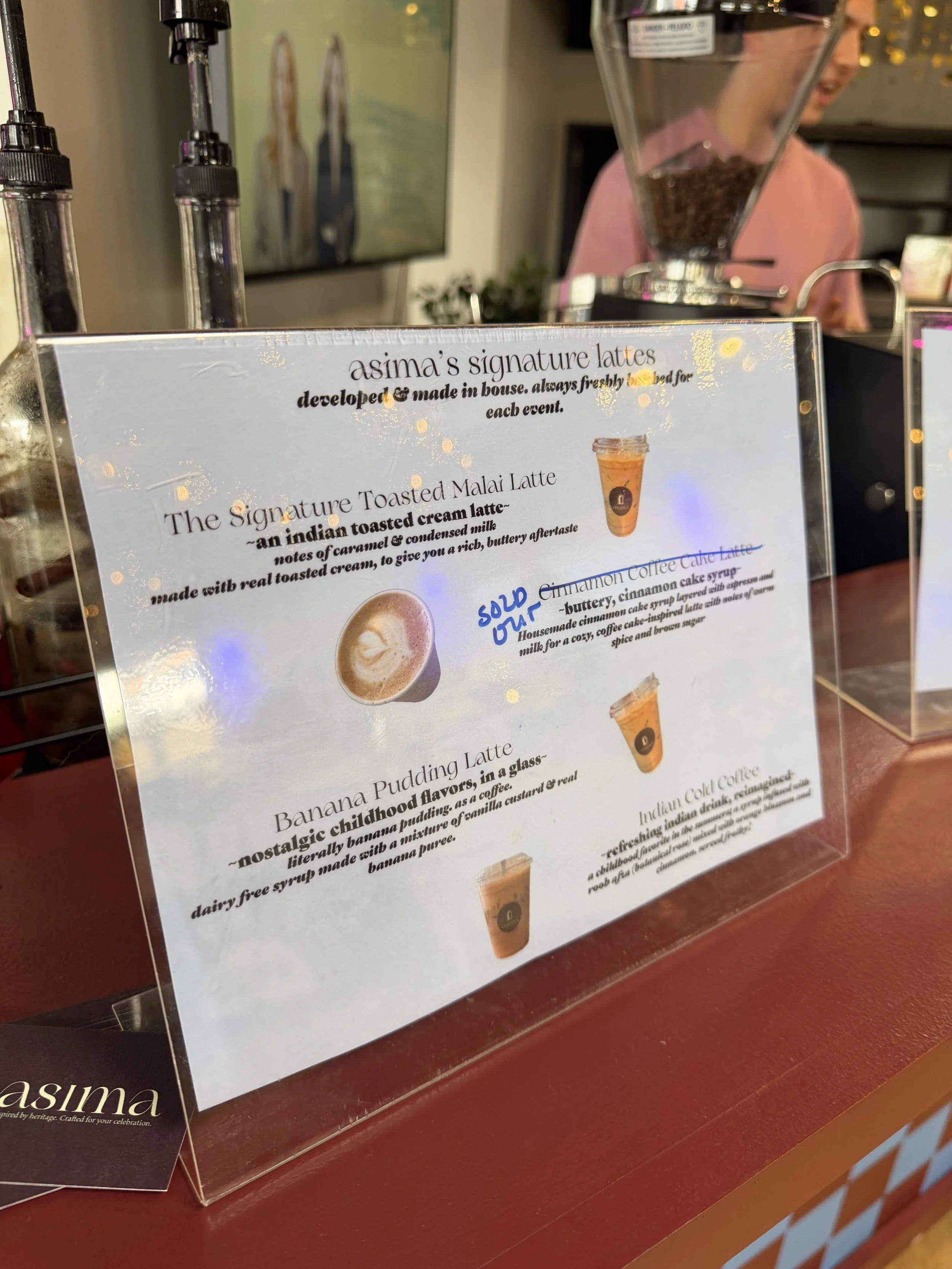 Close-up of a menu display featuring 'Asima's signature lattes' with descriptions of various latte options, including a toasted Malai latte and banana pudding latte, on a red counter.