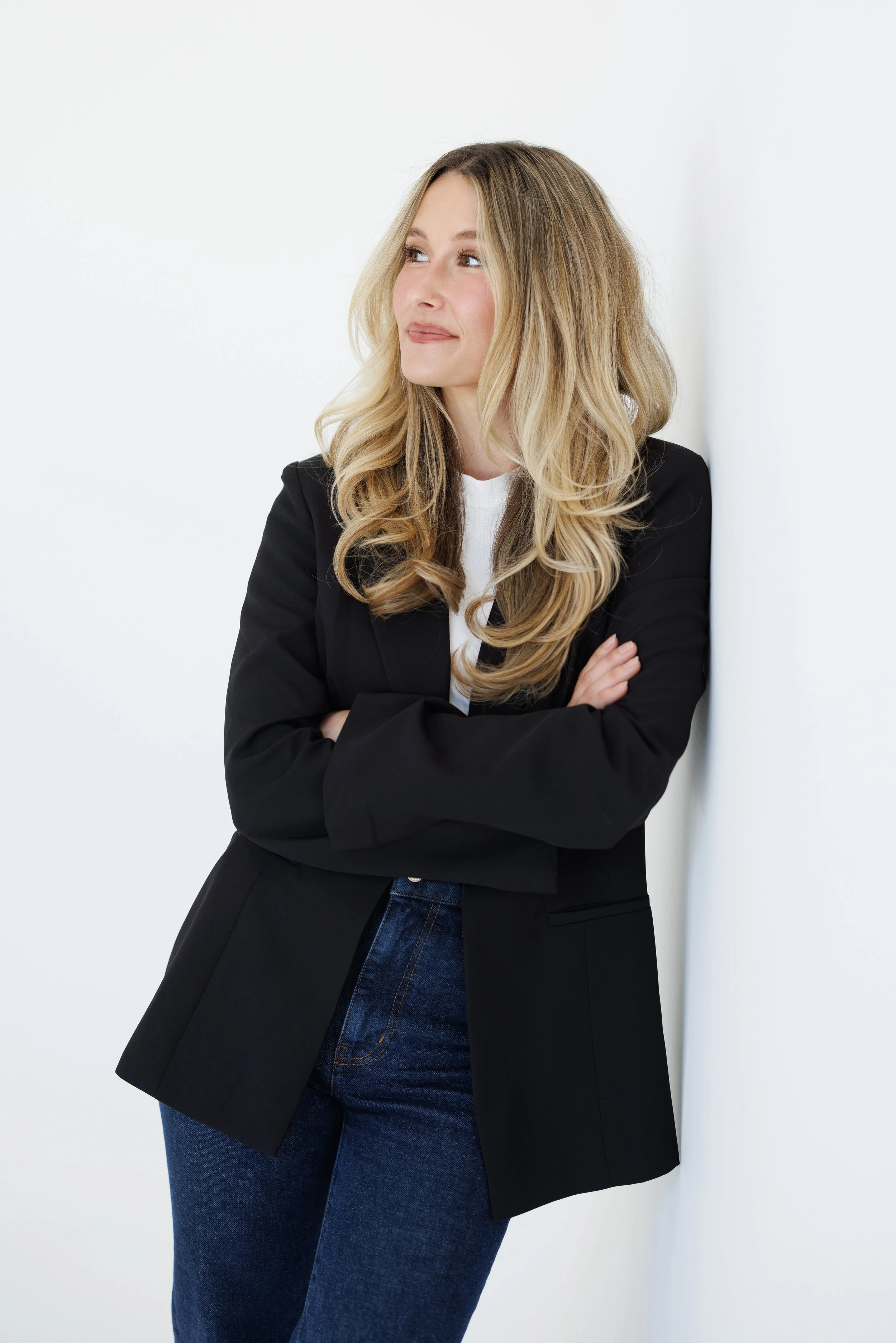 A woman with long wavy blonde hair and light skin, wearing a black blazer over a white shirt, and blue jeans, standing against a plain white wall with her arms crossed, looking slightly to the side.