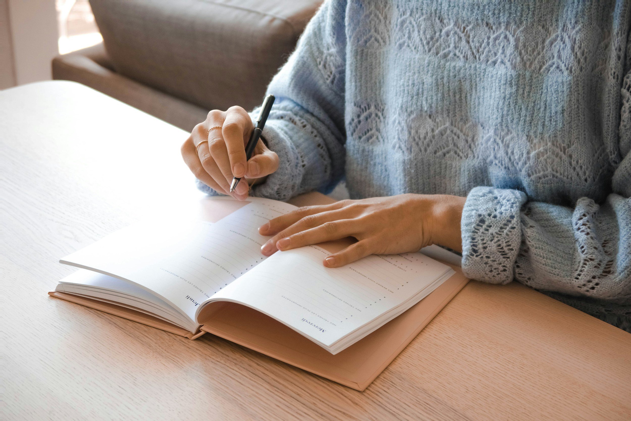 Person writing in a planner or journal at a wooden table, wearing a light blue, lacy knit sweater.