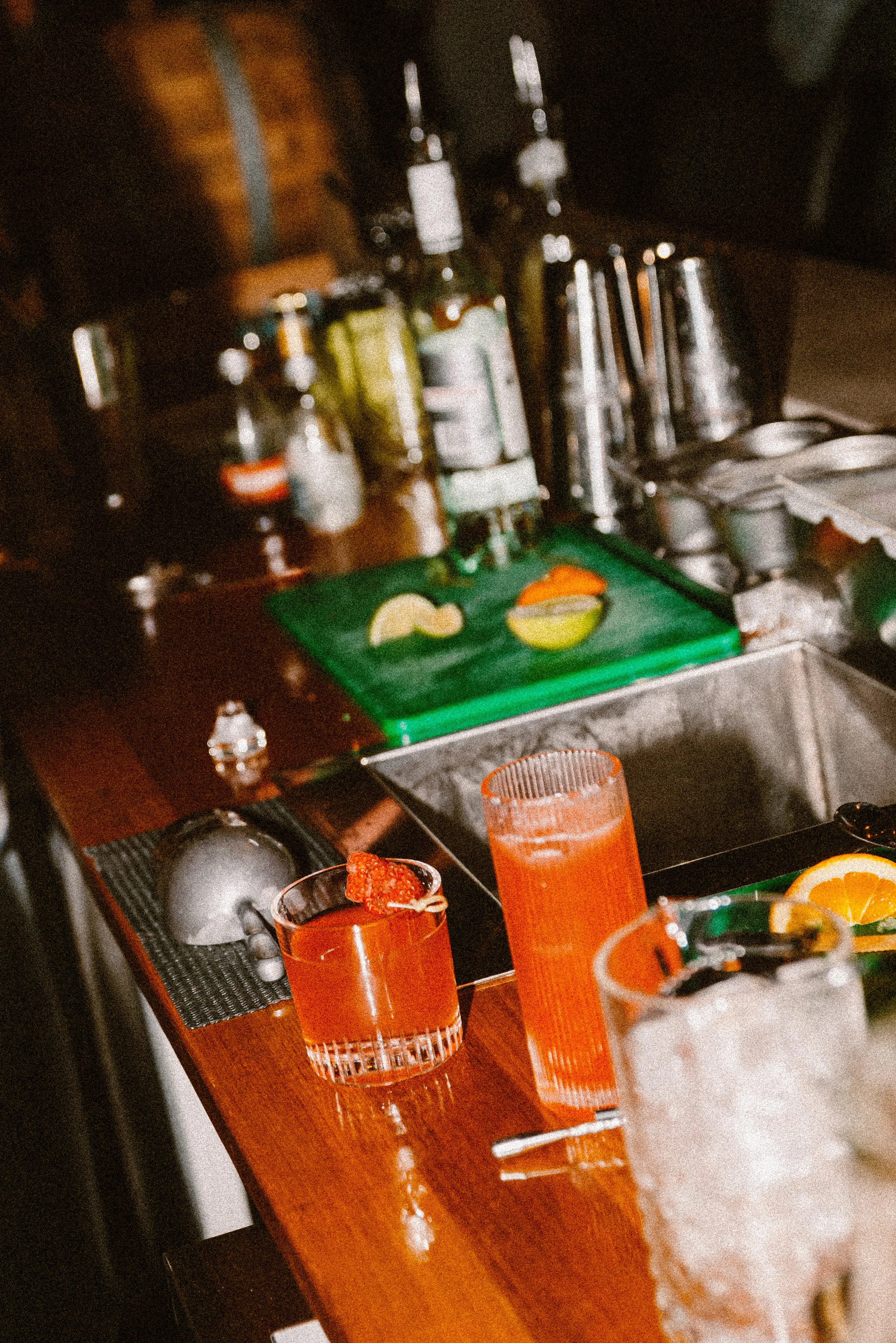 A bar counter with various bottles of liquor, a cutting board with sliced fruit, and two drinks—one with a cherry garnish and another in a tall glass—against a dark background.