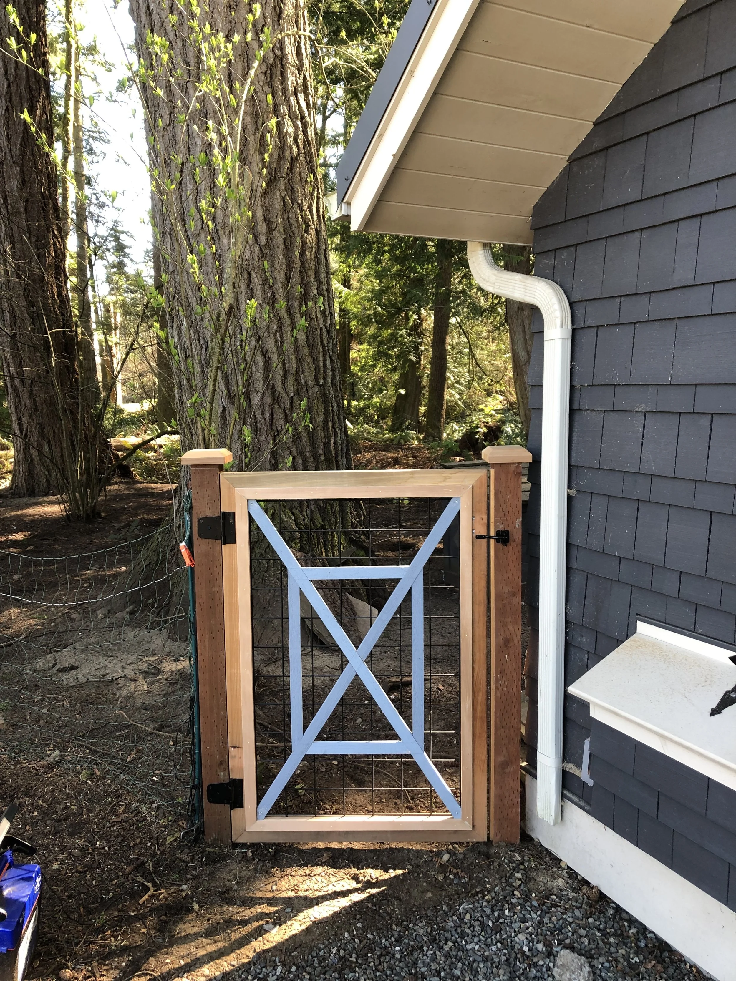 Small gate made of wood and wire mesh next to a house with dark gray siding. Large trees and greenery are in the background.