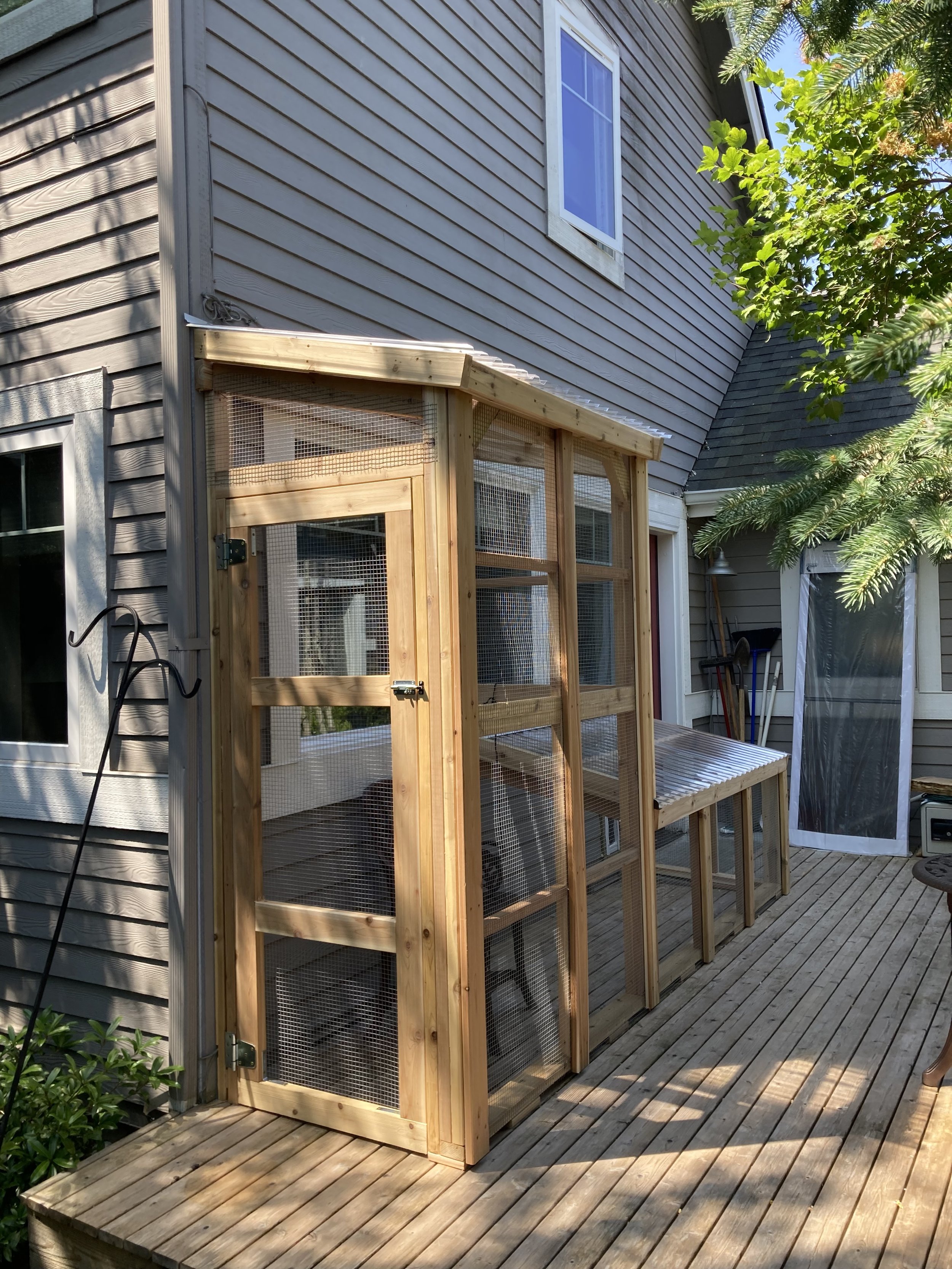 Wooden outdoor chicken coop with mesh wire sides on a wooden deck next to a house with gray siding, surrounded by green trees.