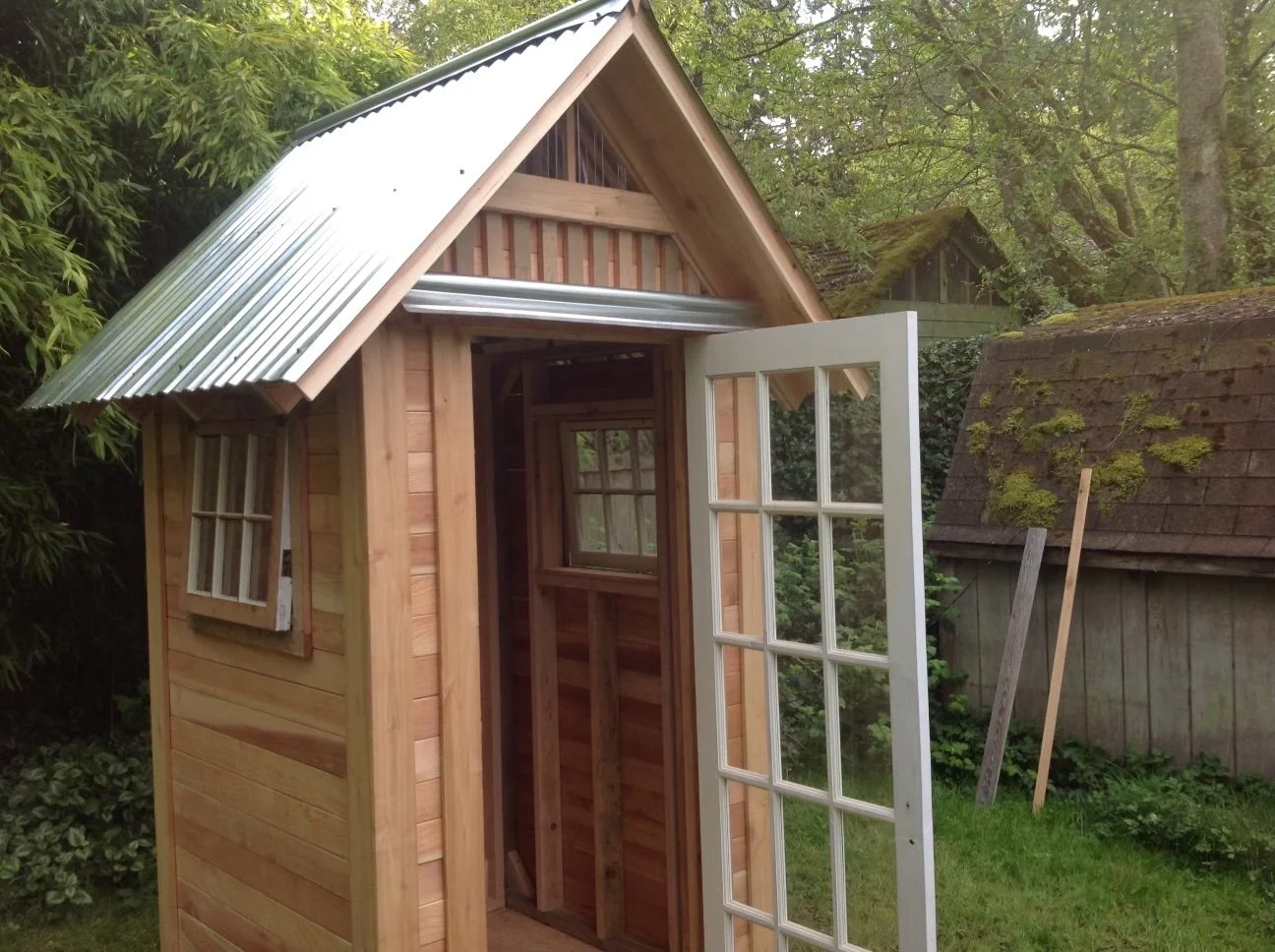 A small wooden shed under construction with a metal roof, a window, and an open door, situated in a lush, green garden.