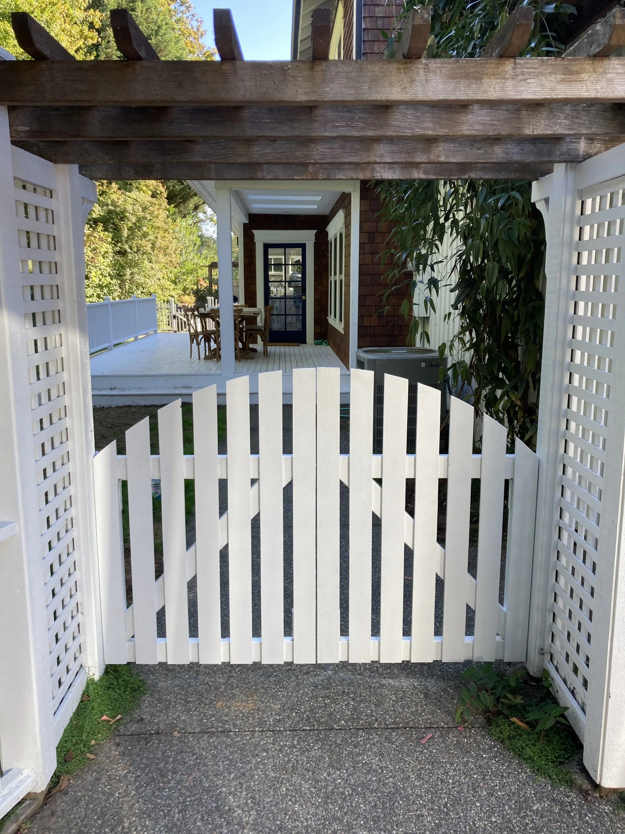 View through a white picket fence gate onto a wooden deck with outdoor dining table, chairs, and a door of a house with large windows.