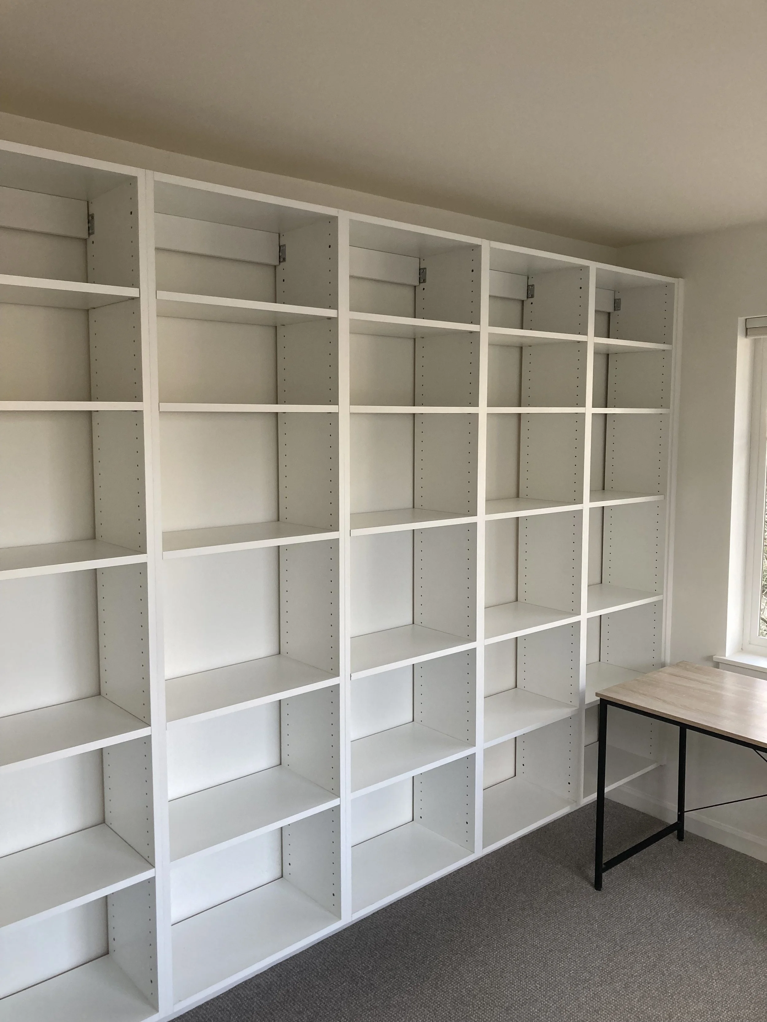 Empty white bookshelves in a room with a window and a small wooden table.