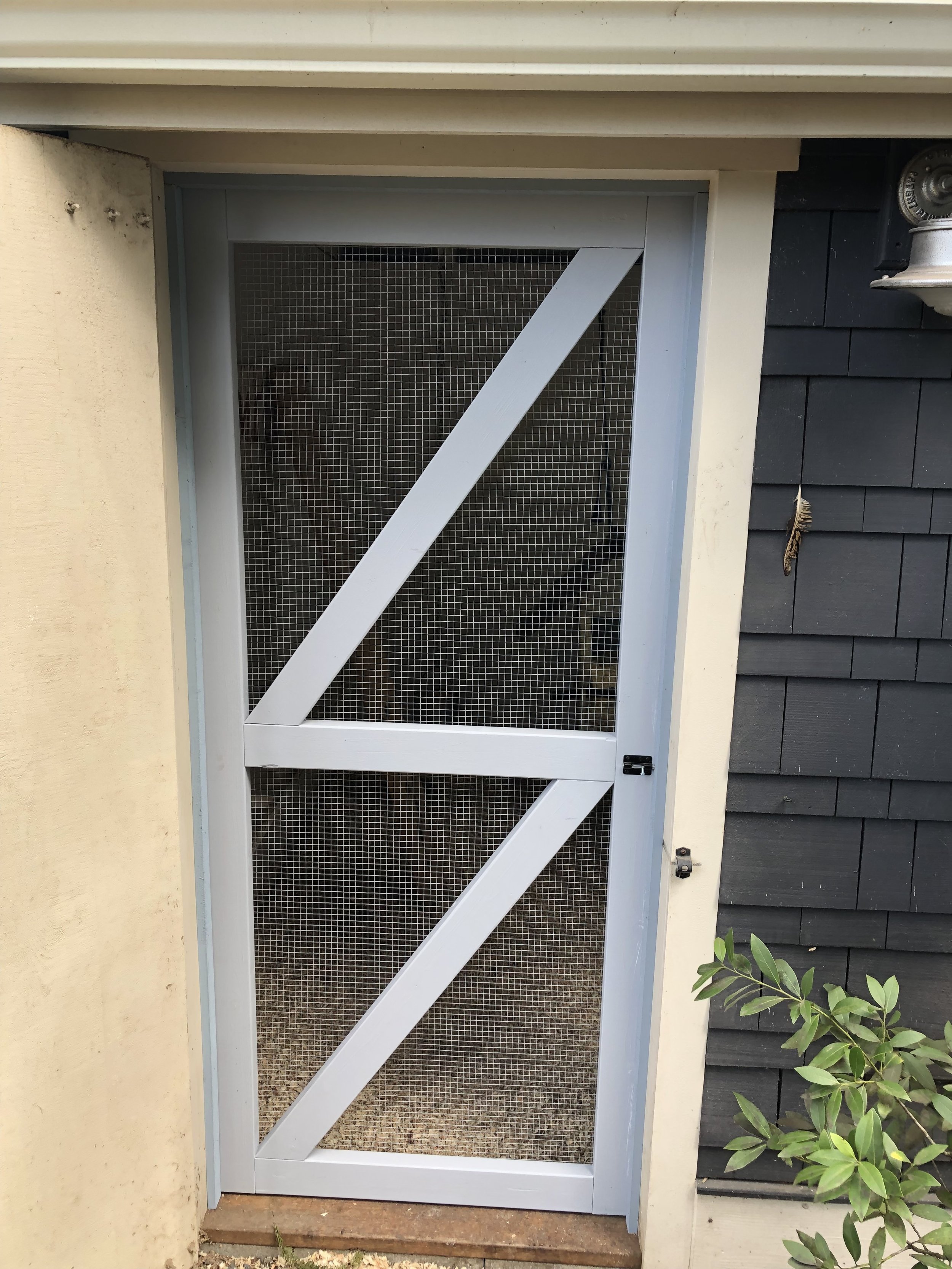 White screen door with wire mesh, installed in a door frame between a beige wall and black shingle siding, with a small green plant in front.