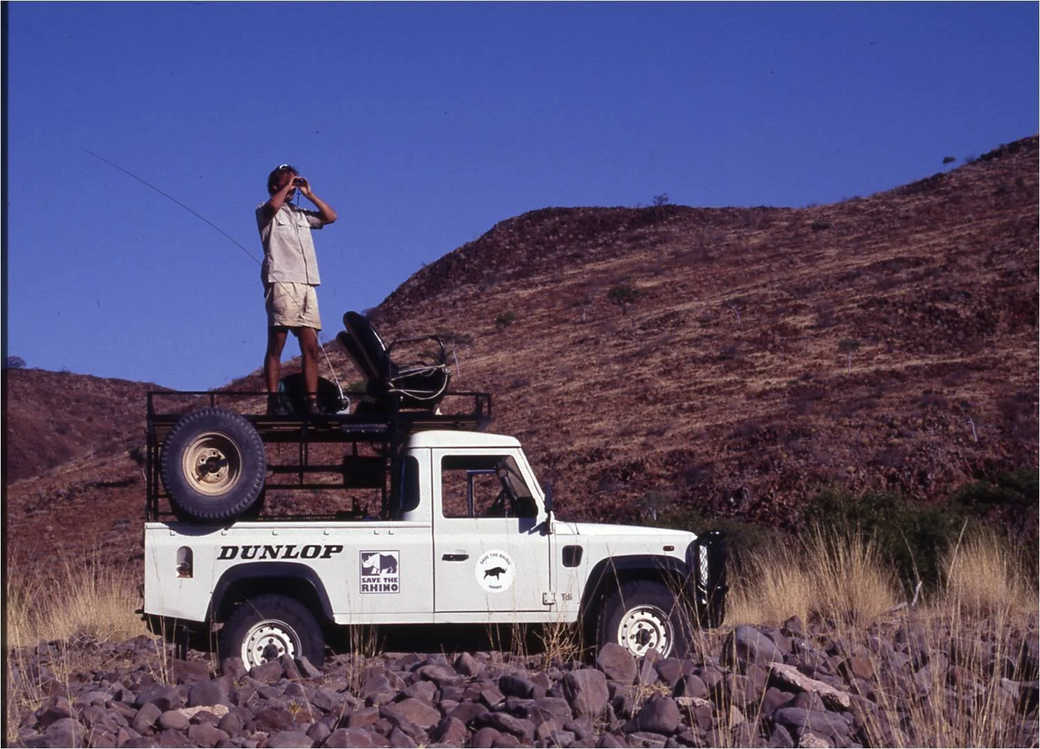 A person standing on the back of a white vehicle with 'DUNLOP' and 'SAVE THE RHINO' stickers, using binoculars to look into the distance in a rocky, grassland landscape with brown hills under a clear blue sky.