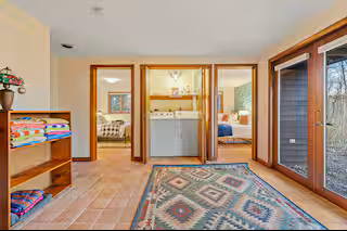 View of a living area with a sliding glass door leading outside, a colorful rug, and a closet with two sliding doors showing bedrooms and a bathroom behind. A wooden shelf with folded towels and decorations is on the left.
