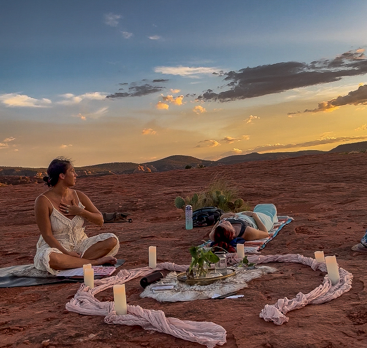 People participating in a meditation session on a desert landscape during sunset, with candles and a decorative cloth on the ground.