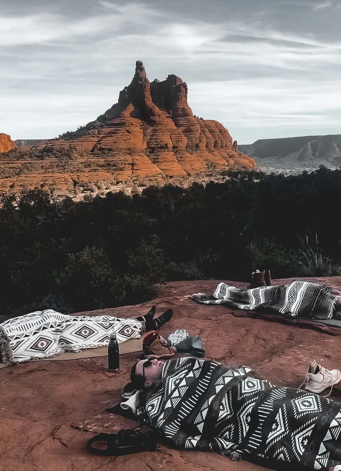 People lying on blankets set up on a rock surface, overlooking a desert landscape with a prominent layered red rock formation in the background, under a cloudy sky.