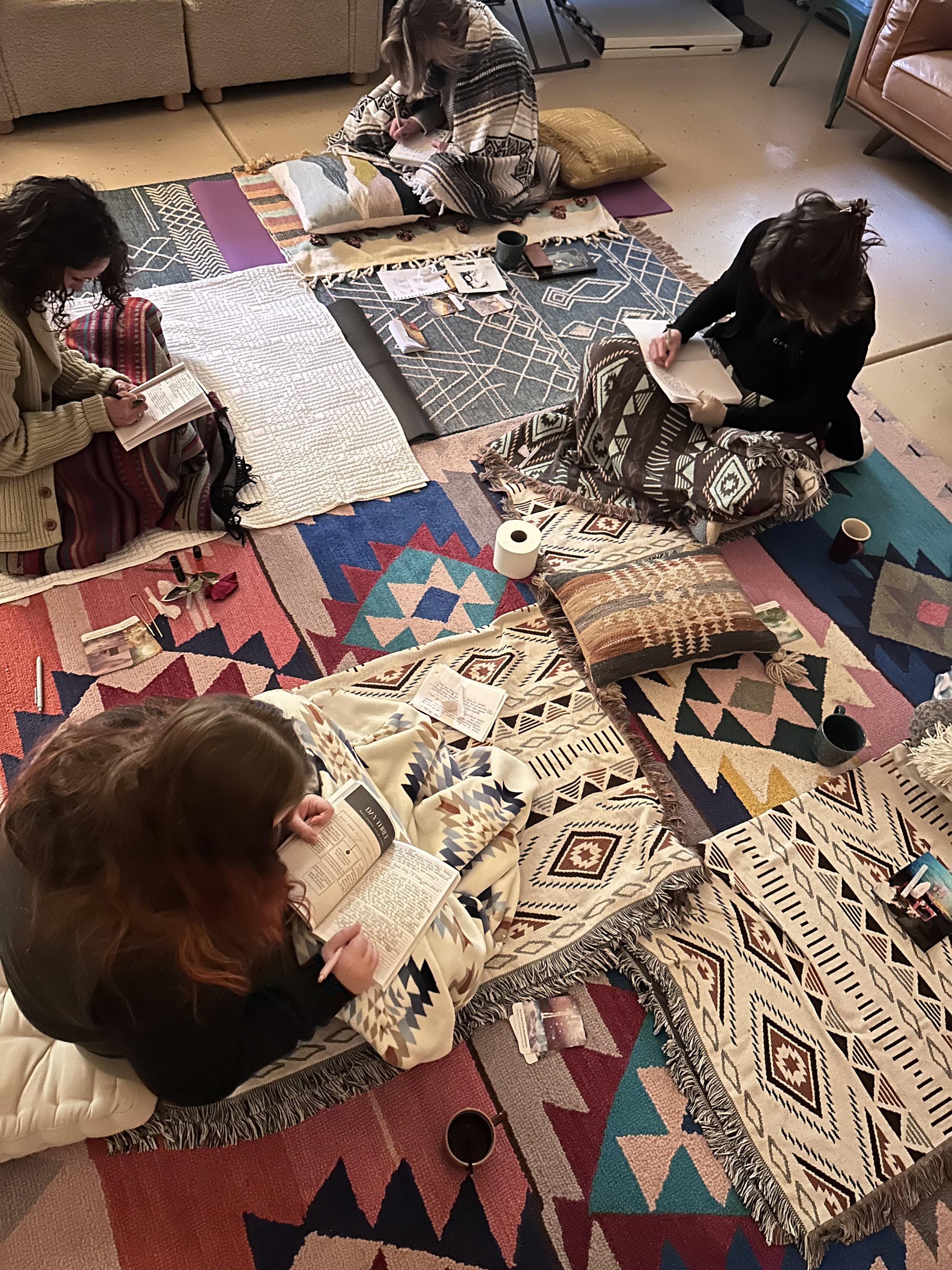 Four women sitting on colorful patterned rugs and cushions on the floor, reading books, with notebooks, cups, and various personal items around them in a cozy indoor setting.