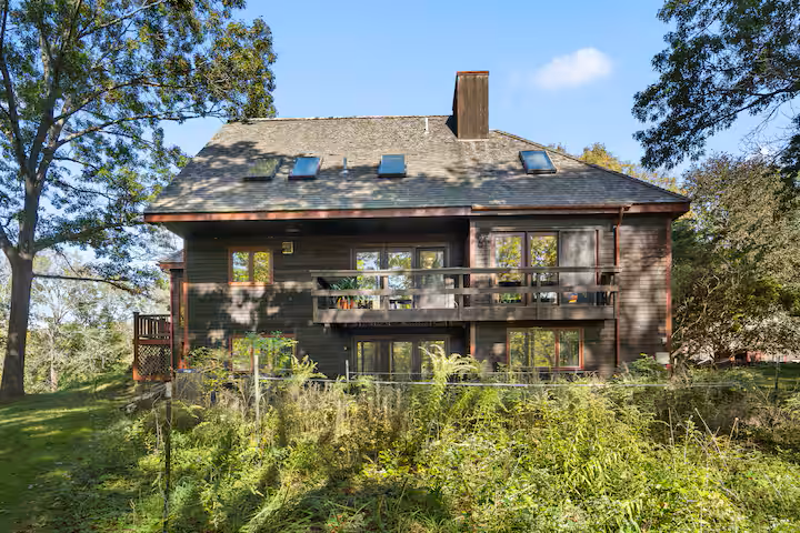 Back view of a two-story wooden house with a balcony, surrounded by trees and greenery, under a partly cloudy sky.