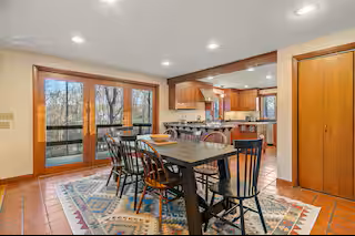 Open kitchen and dining area with large windows, a wooden dining table with chairs, and a colorful rug.
