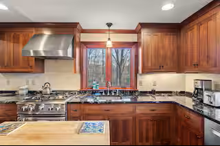 A kitchen with wooden cabinets, a central window, stainless steel range stove, overhead vent, and various small appliances on the countertops.