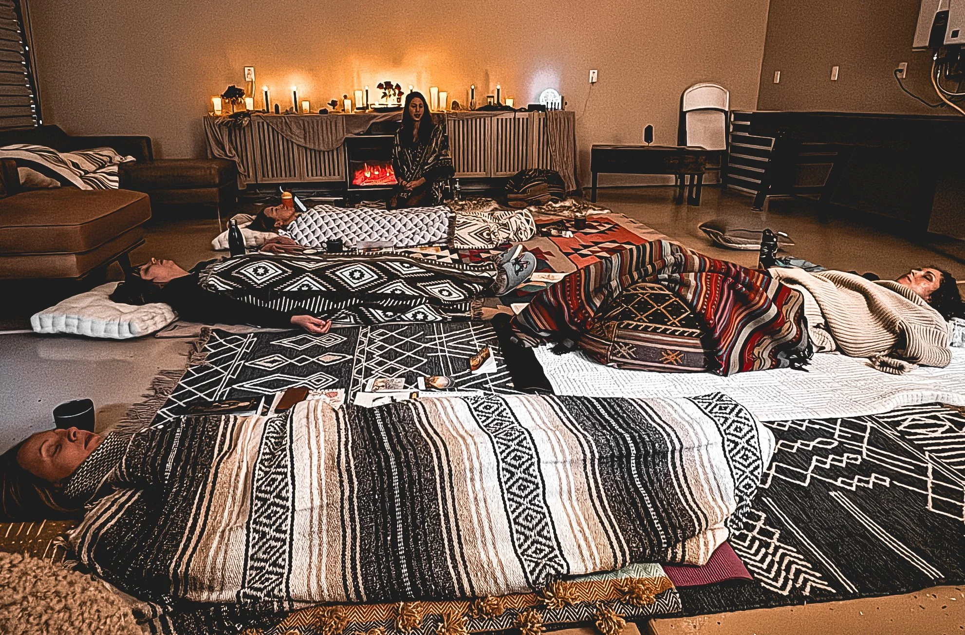 People lying on the floor on blankets and pillows during a cozy gathering in a dimly lit living room with candles and a fireplace.