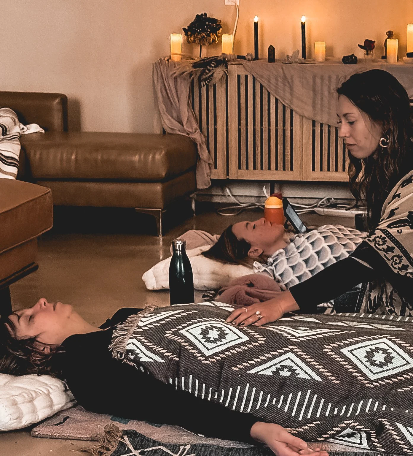 Three women lying on the floor with candles and decorations in the background. One woman is resting on a pillow with a blanket over her, while another woman is lying on her side, looking at a phone, and the third woman is kneeling beside her, placing a hand on her.