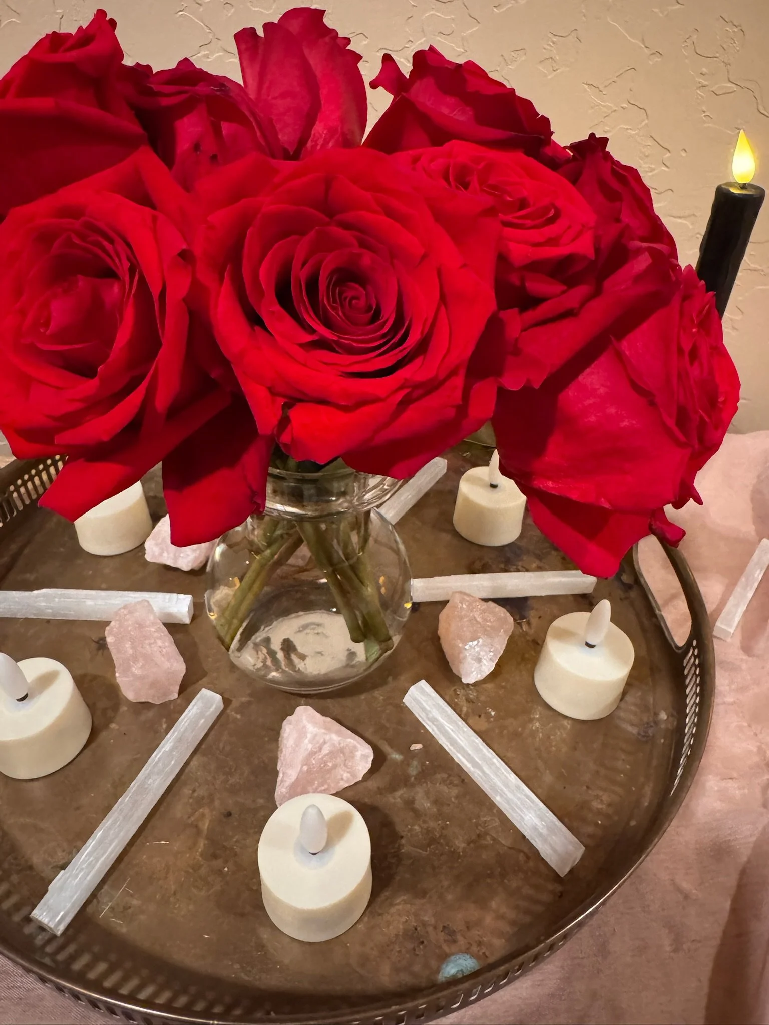 A bouquet of red roses in a glass vase on a tray decorated with white candles, pink rocks, and white sticks.