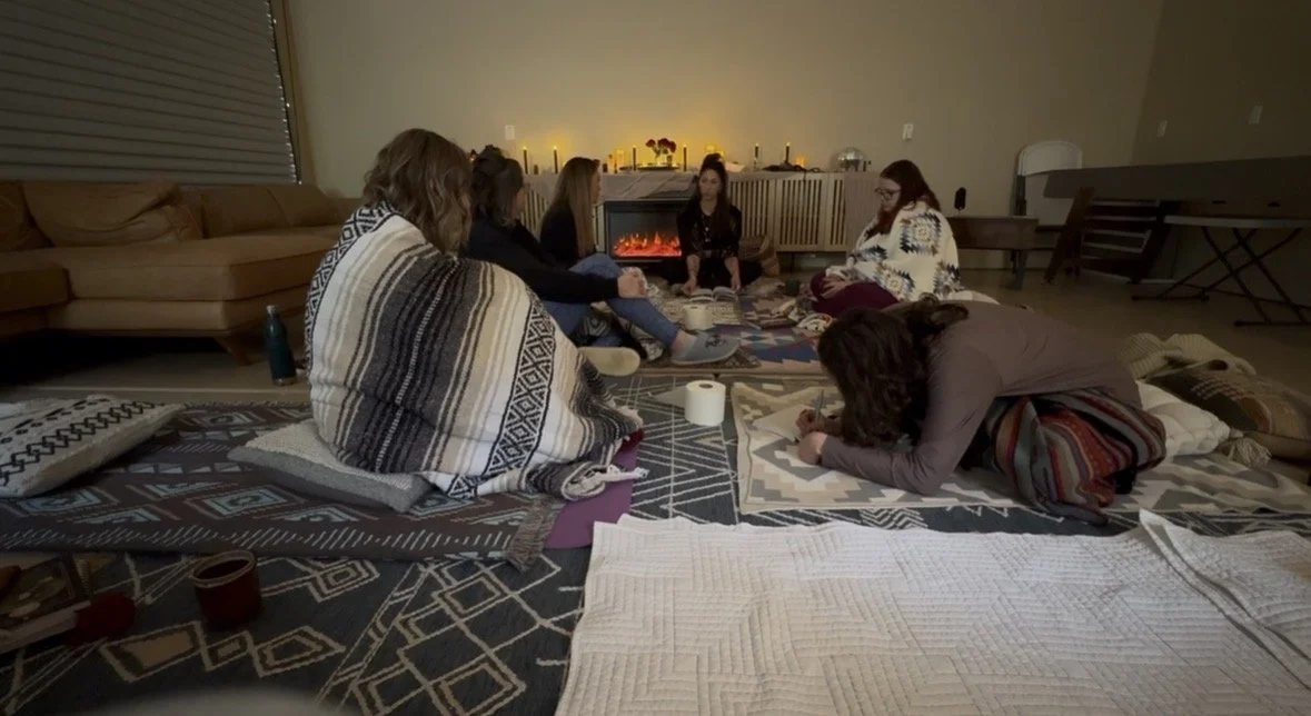 A group of people sitting on the floor in a living room around a table playing a card game, with a fireplace and candles in the background.