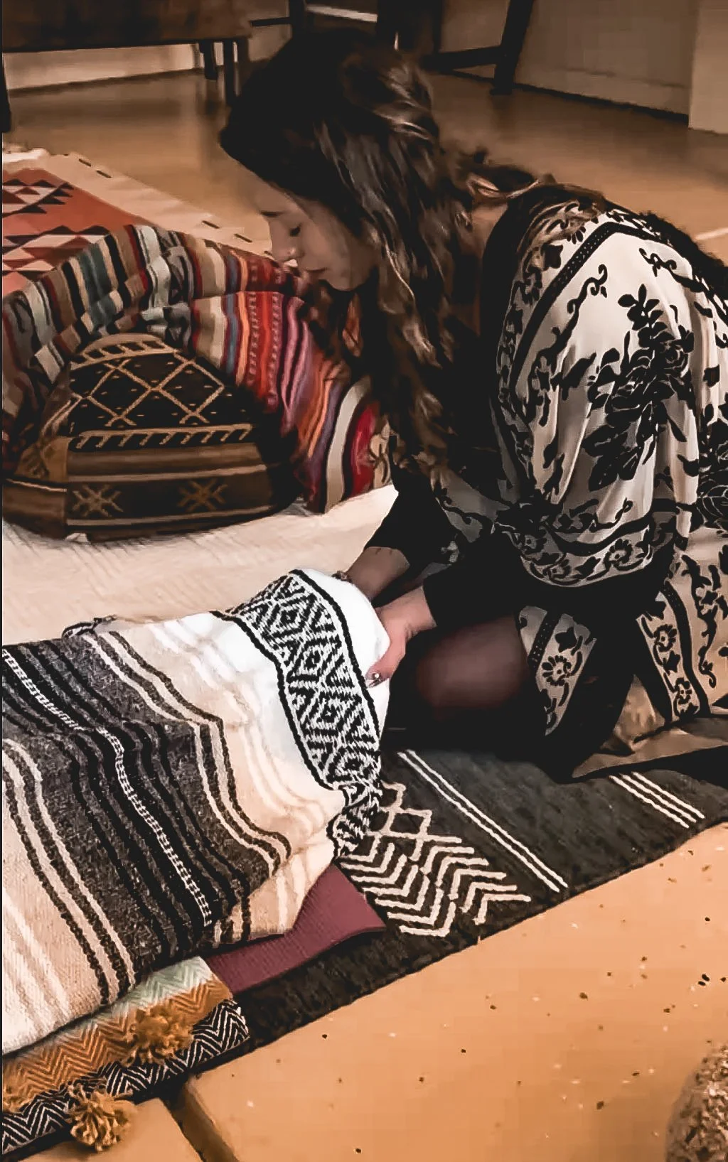 A woman with shoulder-length brown hair is kneeling, holding and comforting a person wrapped in a black and white patterned blanket. They are on a geometric patterned rug in a room with wooden flooring and a bed with colorful, striped blankets.